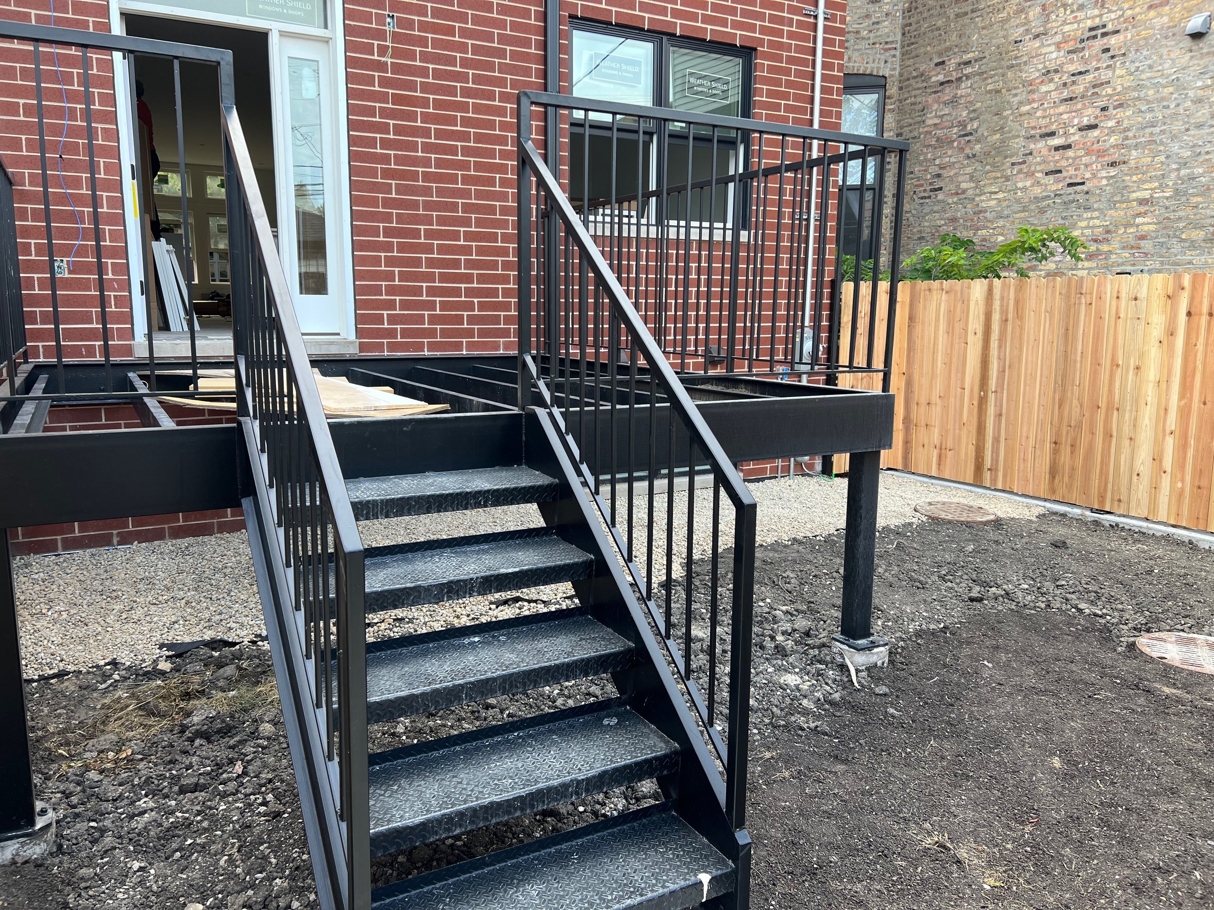 Back door with stairs leading up to a small balcony in a newly constructed residential building. The building exterior combines red brick and a section of weathered brick. There's a wooden fence on the right side and construction materials on the gro