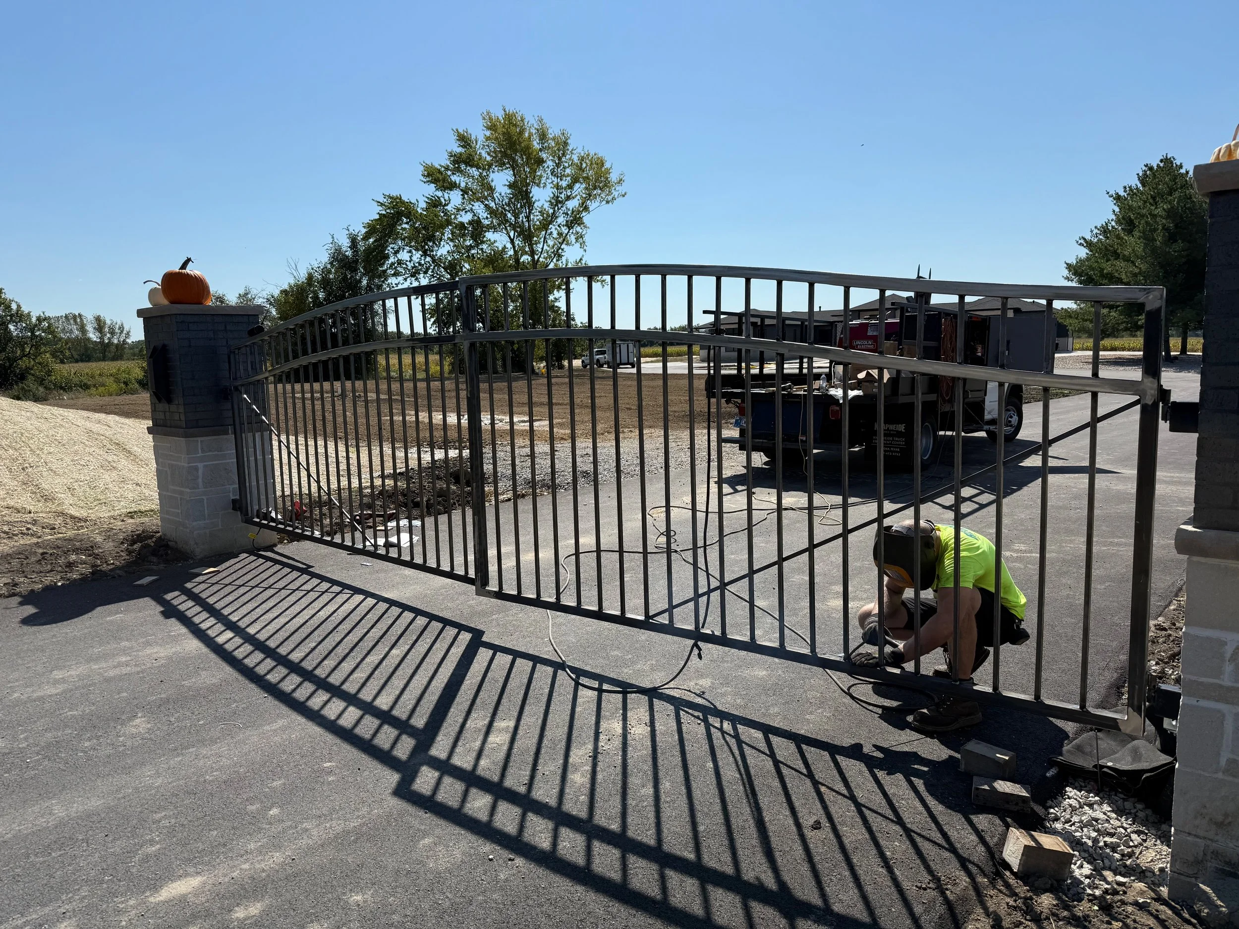 A worker in a yellow safety shirt installing or repairing a metal gate at the entrance of a property. The gate is attached to brick pillars topped with pumpkins, indicating an autumn or Halloween theme. There are trees and a clear blue sky in the bac