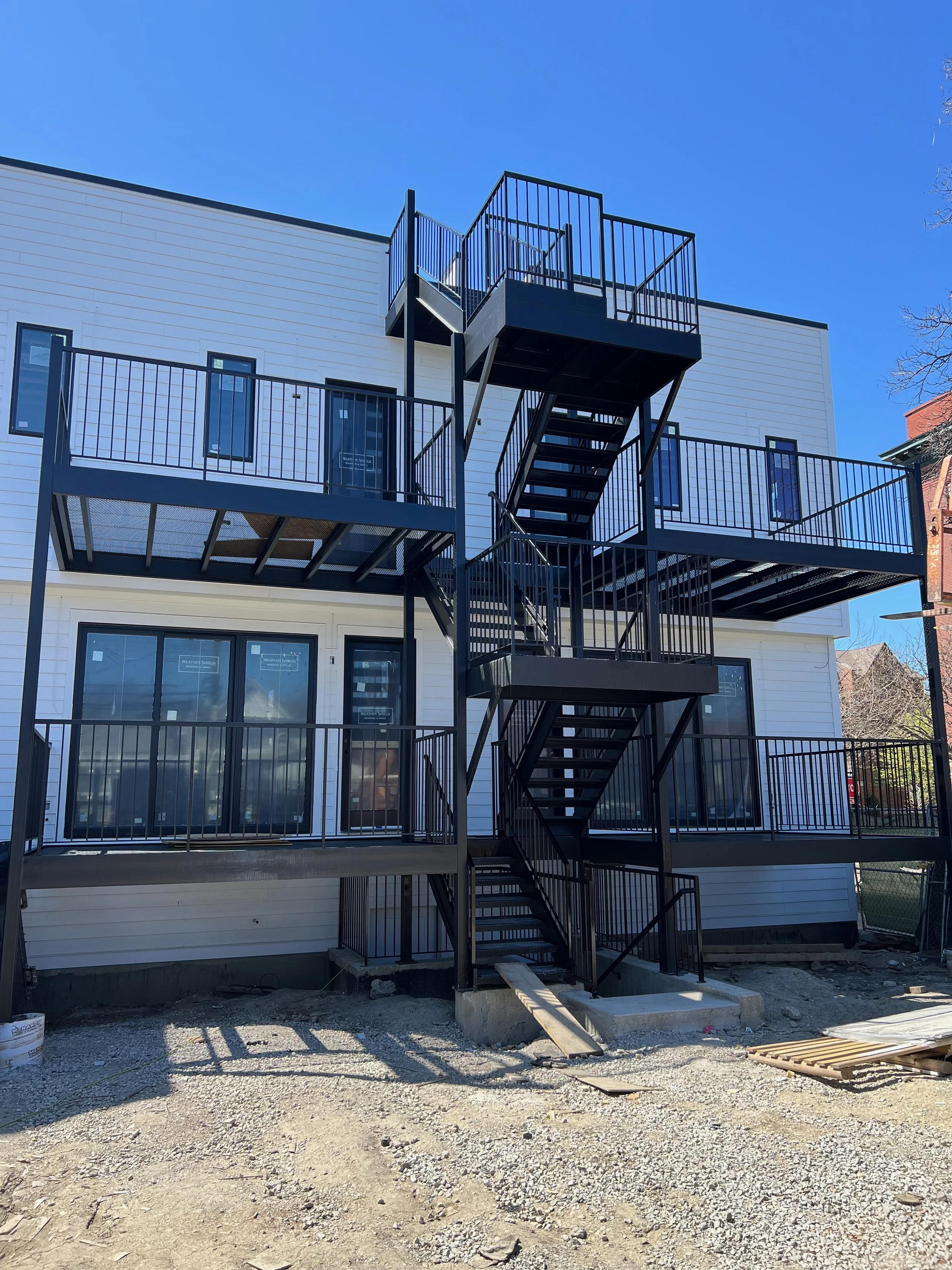 Multi-story residential building under construction with black metal fire escape and balconies, white siding, and large windows, on a clear sunny day.