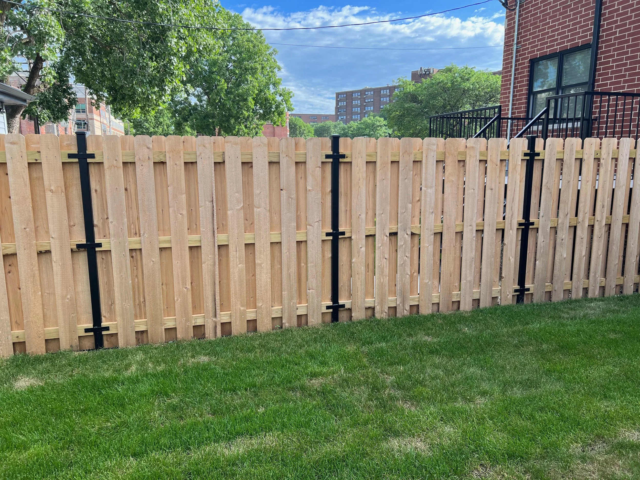 A backyard with green grass, a wooden fence, trees, a brick building with a staircase, and a multi-story residential building in the background under a partly cloudy blue sky.