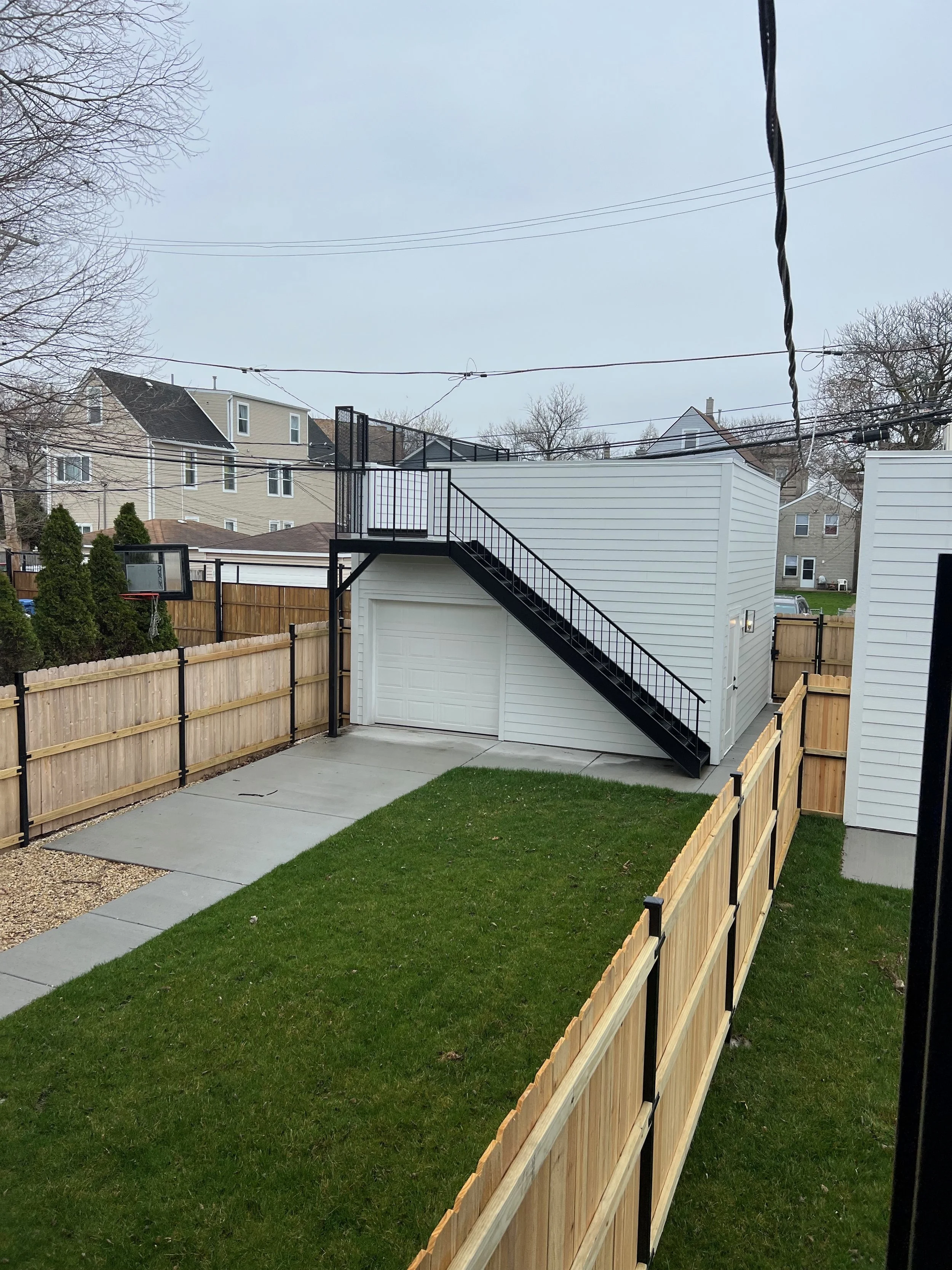 Backyard with a wooden fence, concrete walkway, grass lawn, and a white garage with black stairs leading to a balcony. Houses and trees in the background, overcast sky.
