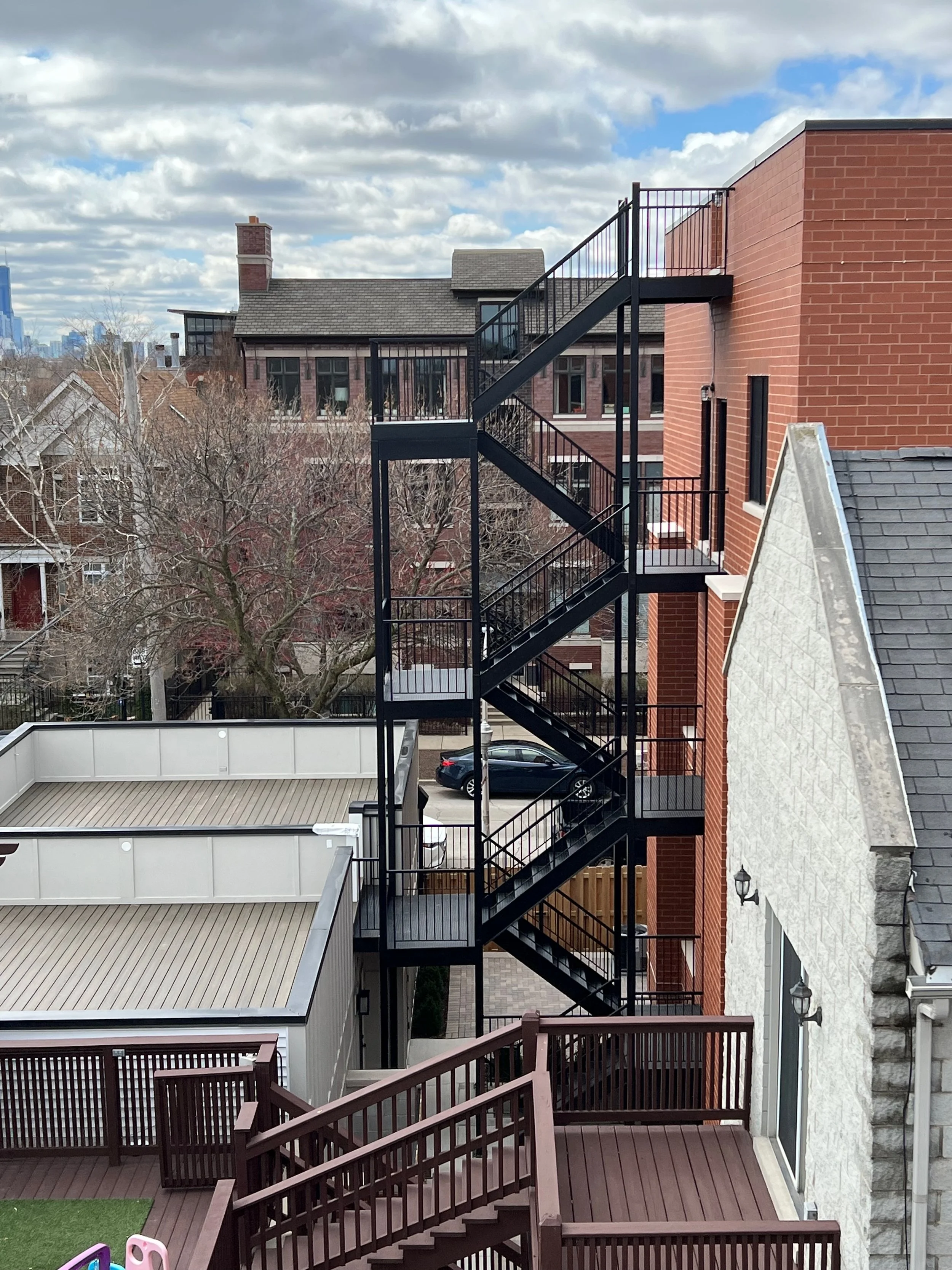 Multi-story residential building with exterior staircase, brick and stone walls, large windows, and a small backyard with a wooden deck and plastic play structures.
