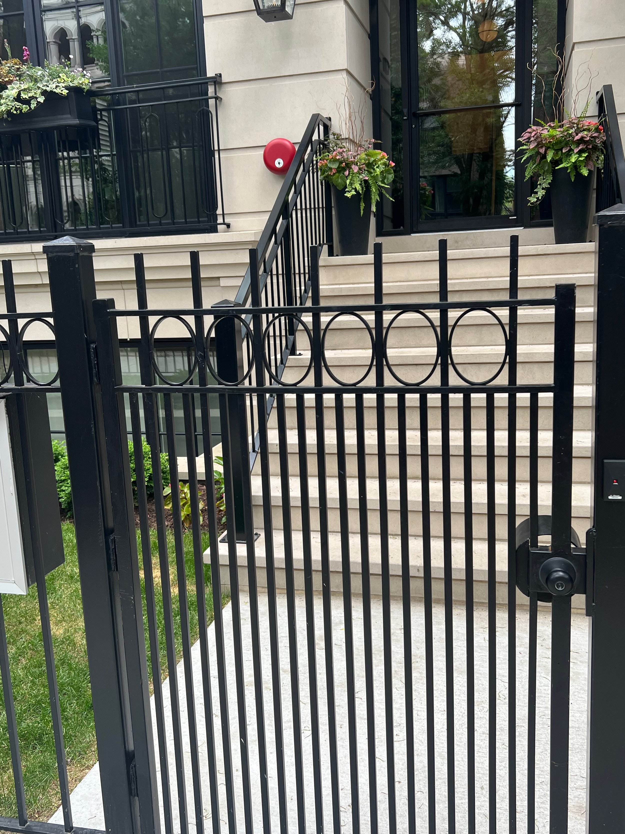 View of a building entrance with stairs, glass door, plants in large black pots, and a black metal fence in the foreground. A red fire alarm box is mounted on the wall near the stairs.
