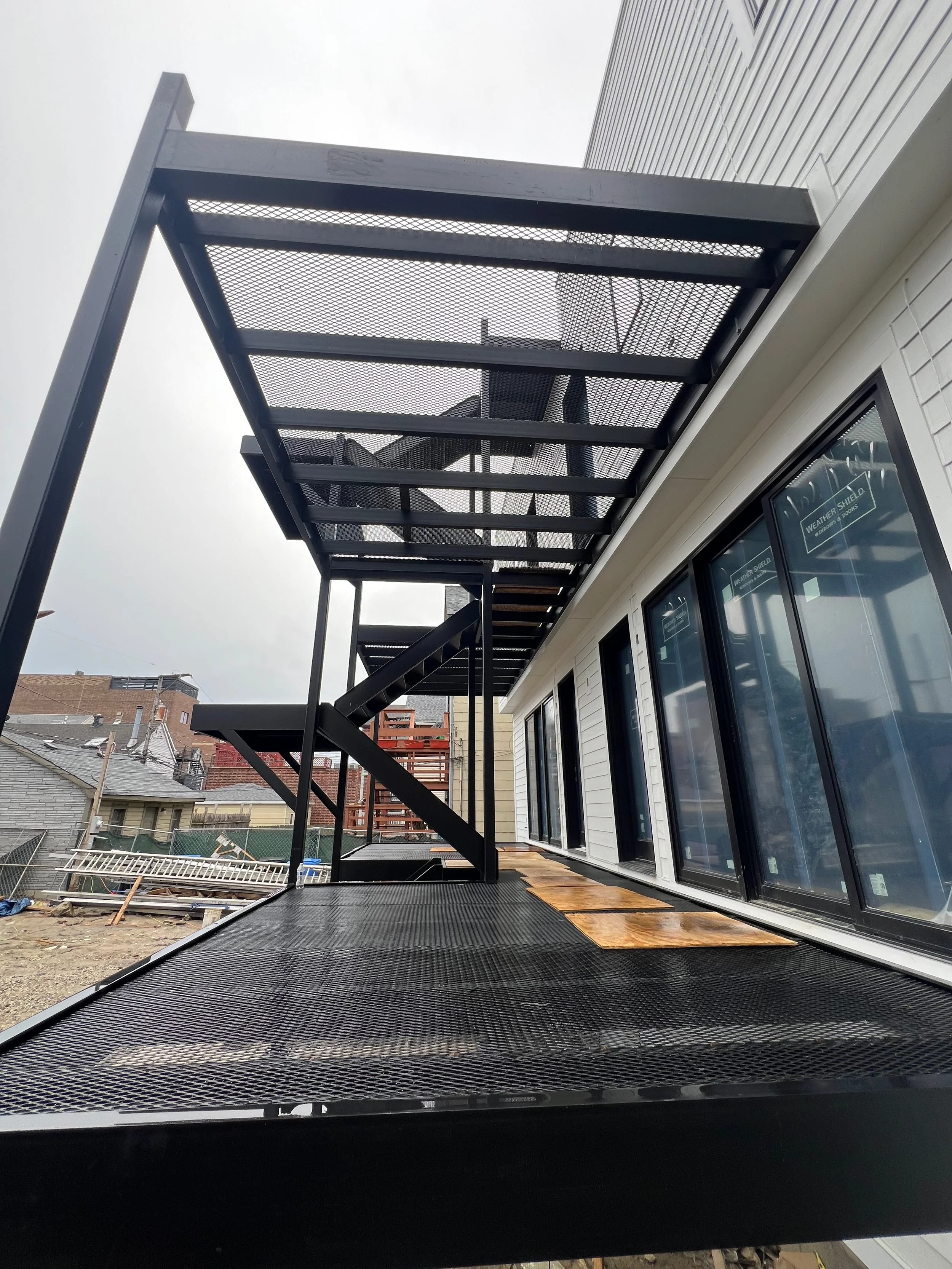 Metal fire escape staircase outside a building with black grating steps and supporting beams, adjacent to large windows.