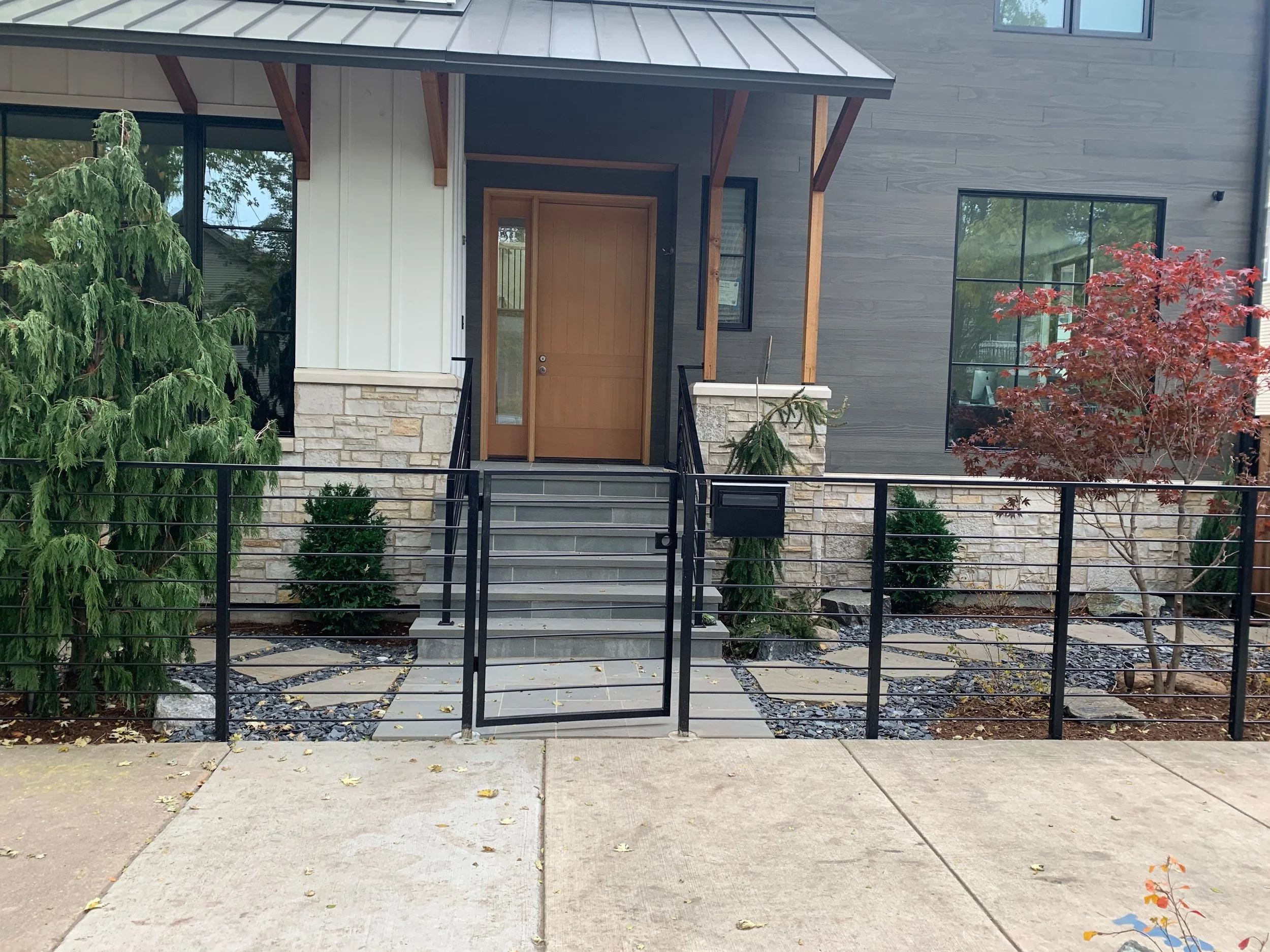 Front view of a modern house with a wooden door, stone steps, black metal railing, and large windows, surrounded by trees with green and red leaves.