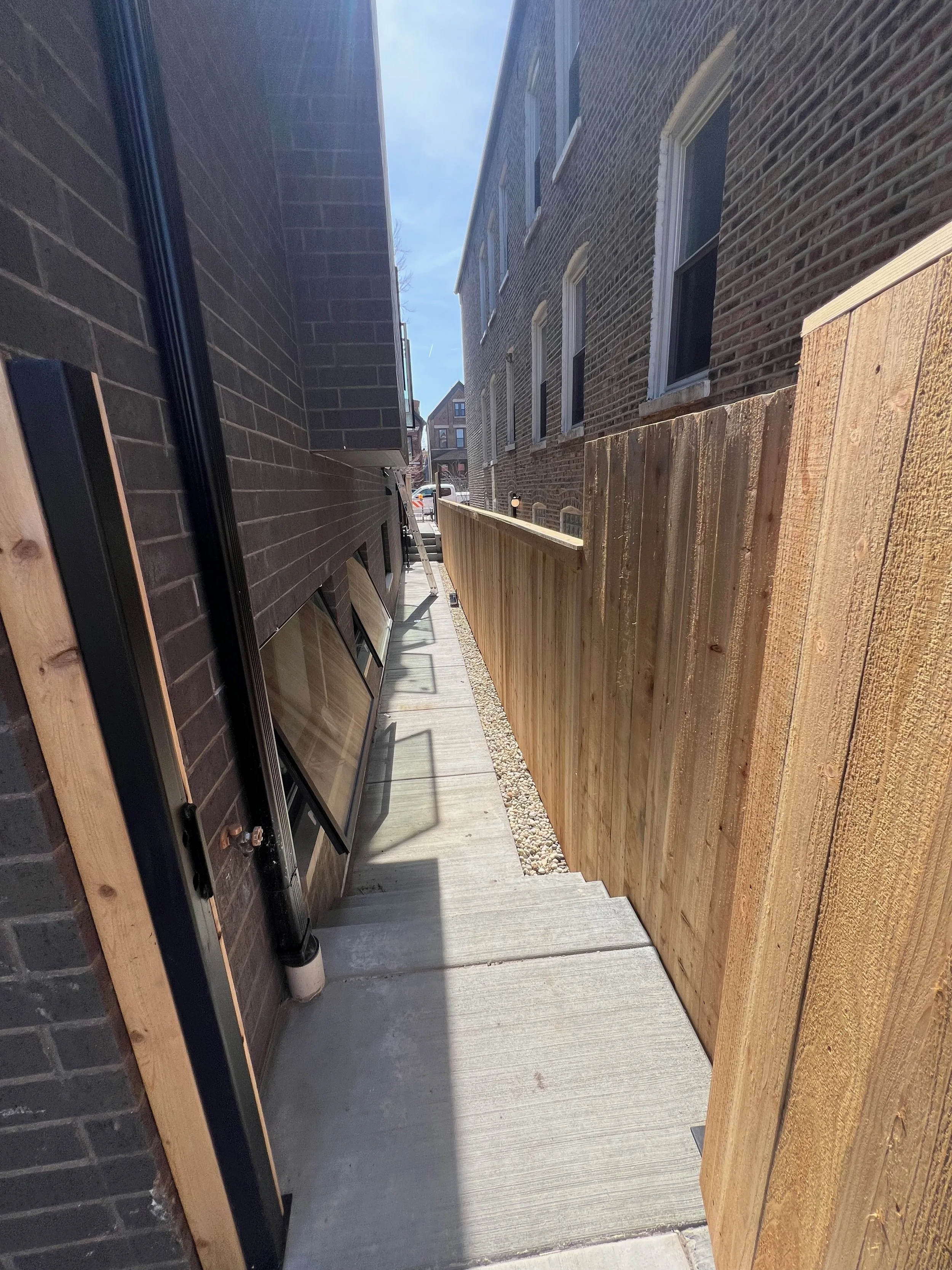 Back alley between brick buildings with stairs and a wooden fence, with a clear sky visible above.
