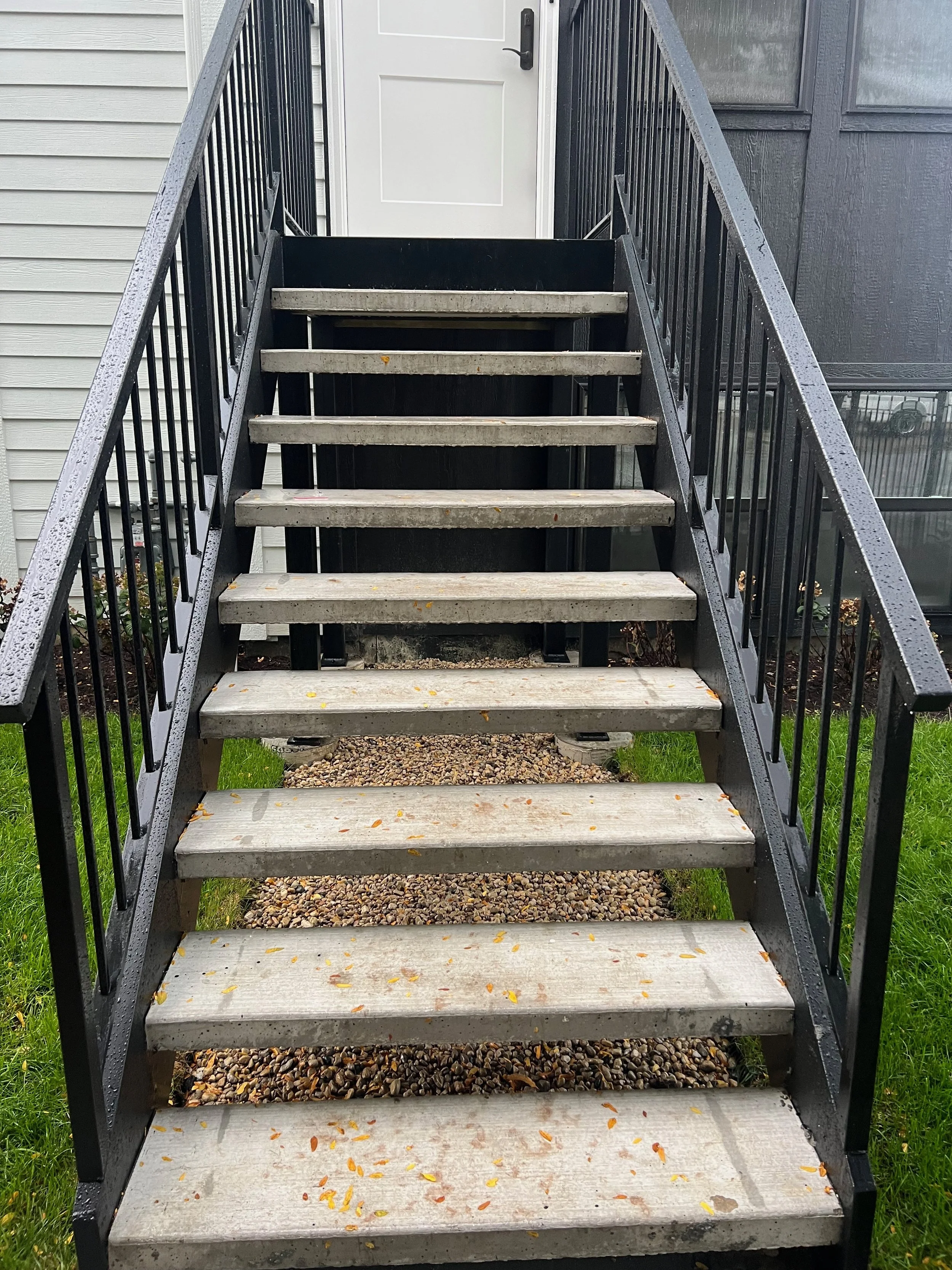 Outdoor metal stairway with raindrops on the handrails and fallen leaves on the steps, leading to a white door with a black door handle, adjacent to a house with gray siding and a screened porch.