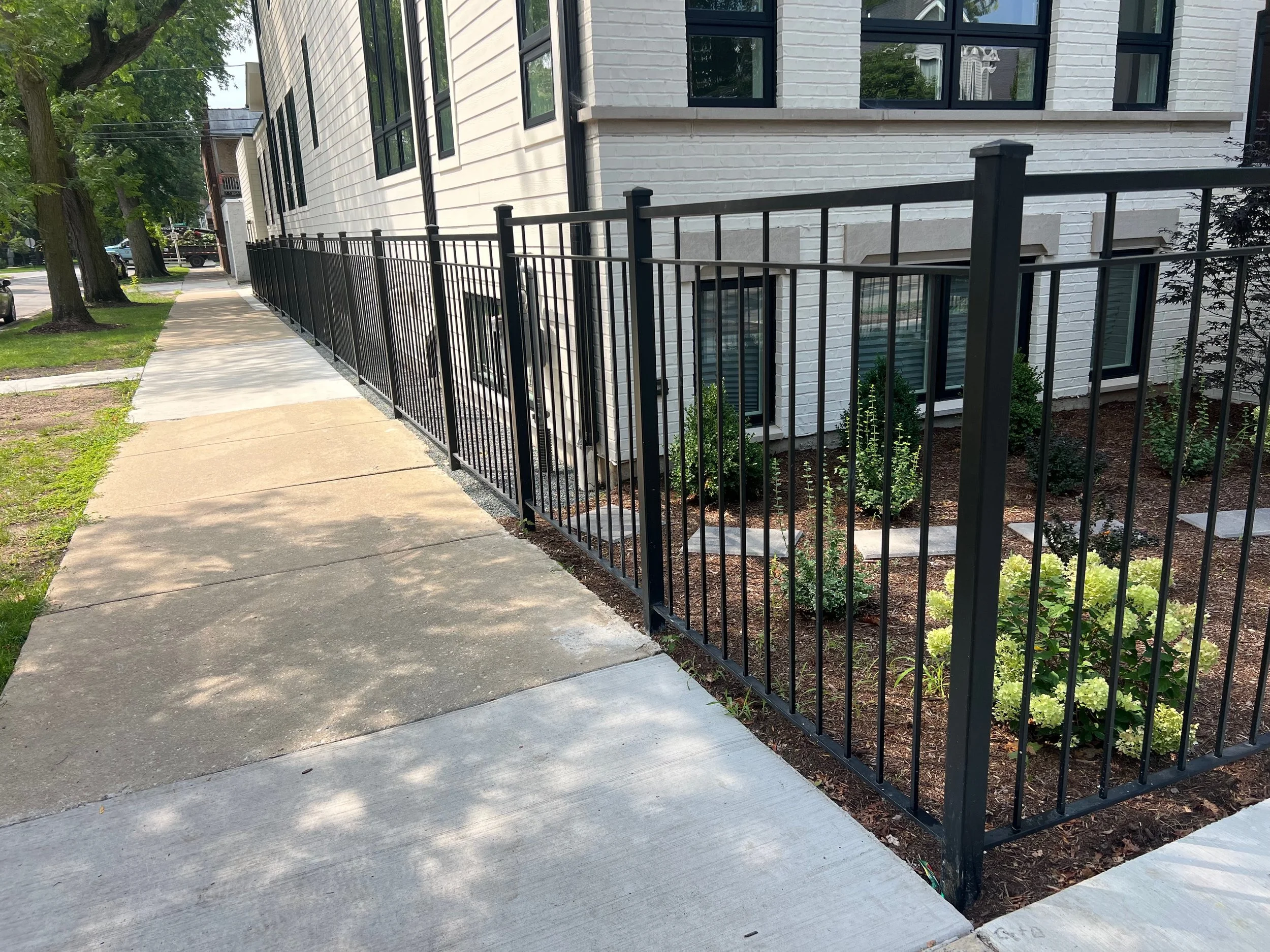 Sidewalk along a building with white brick exterior. Black metal fence runs alongside the sidewalk with some small plants and mulch underneath. Trees and cars are visible in the background.