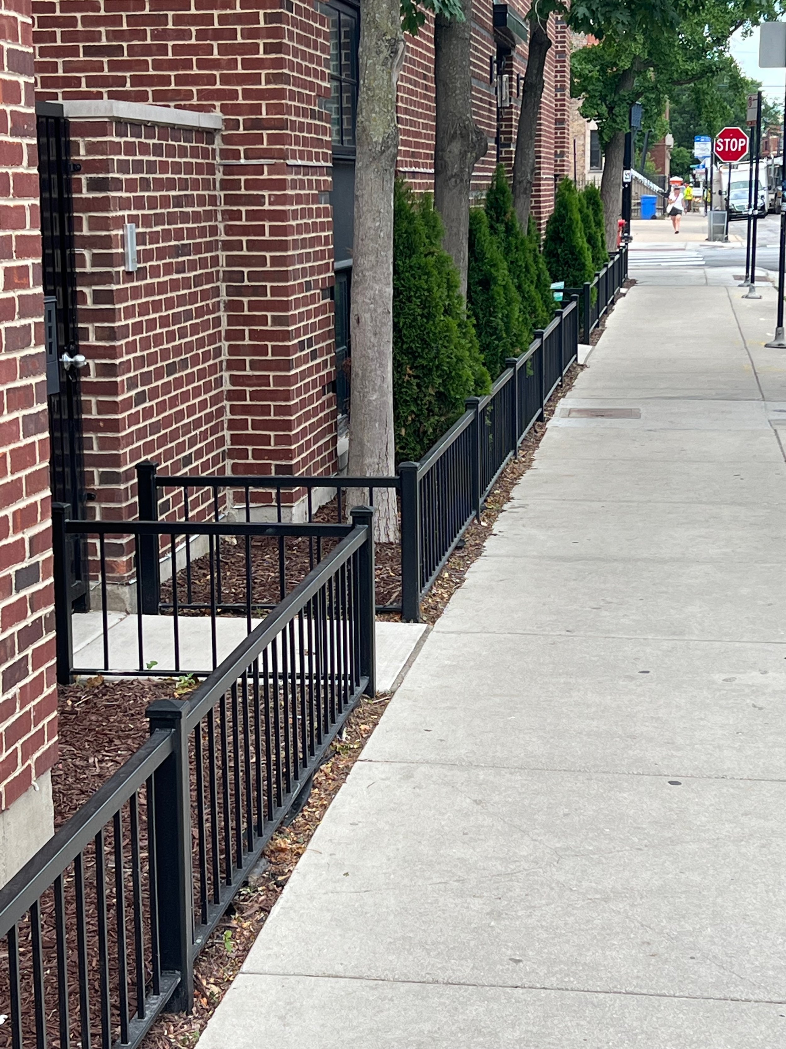 Sidewalk with a row of green bushes, trees, a black metal fence, a red brick building, a stop sign, and some people walking in the background.