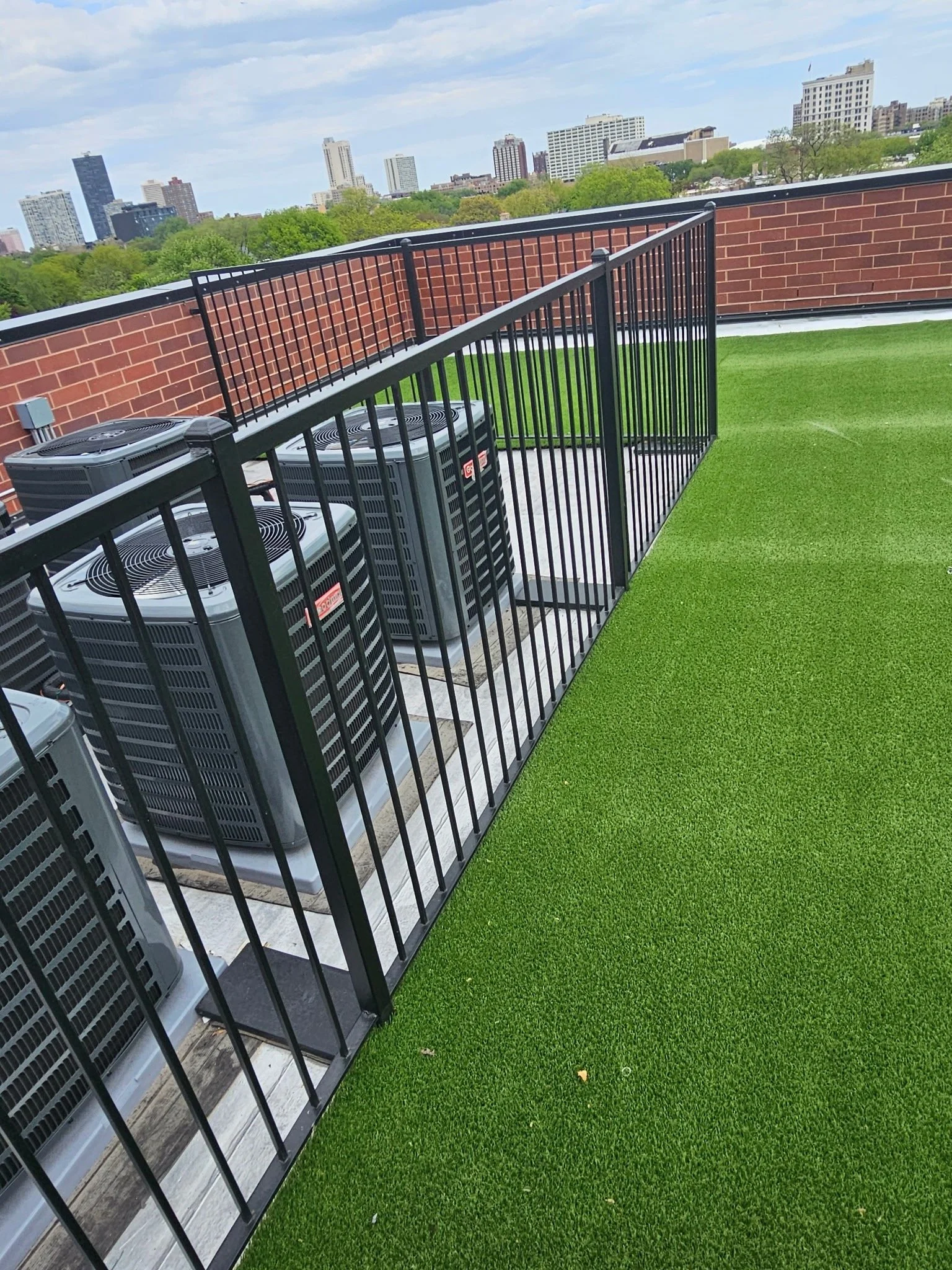 Rooftop terrace with artificial green grass flooring, black metal safety railing, city skyline in the background, and HVAC units on the left.