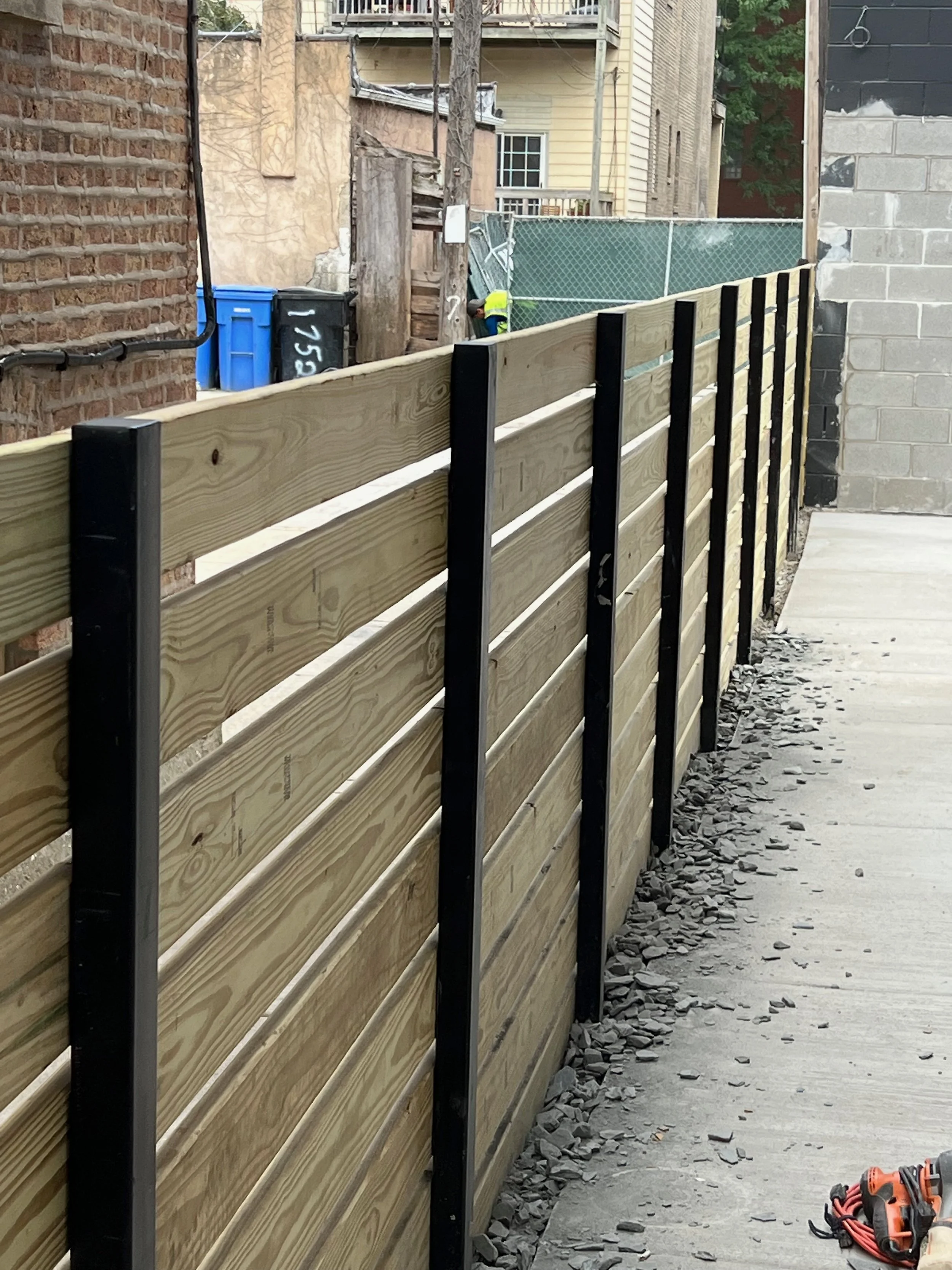 A construction site with a new wooden fence being installed along an alley. The fence has horizontal wooden planks with black metal support posts. There are piles of rocks along the sidewalk, and in the background, there is a person wearing a yellow 