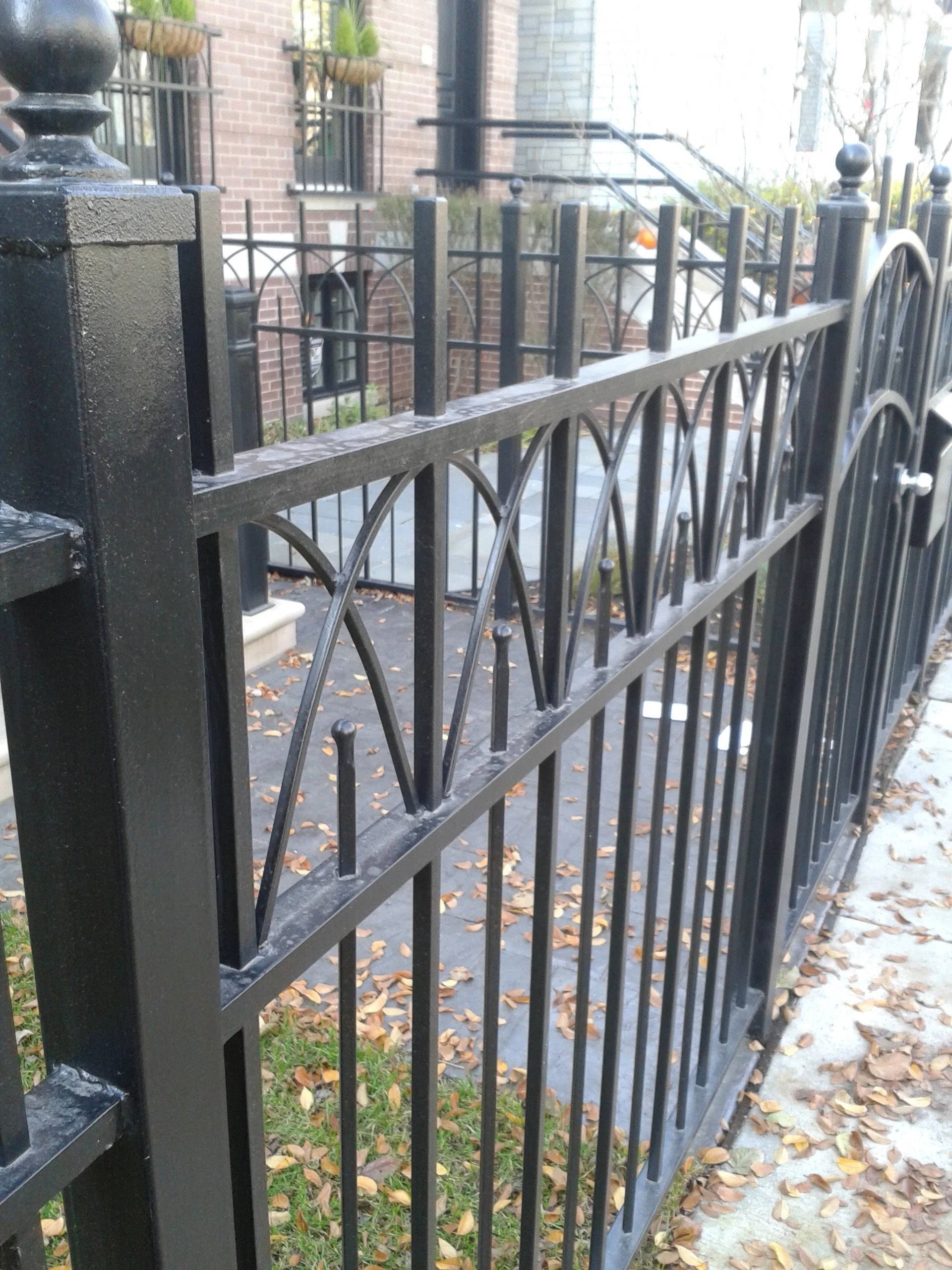 A black metal fence with decorative arches and round finials, located in front of a brick building with windows and potted plants, with fallen leaves on the ground.