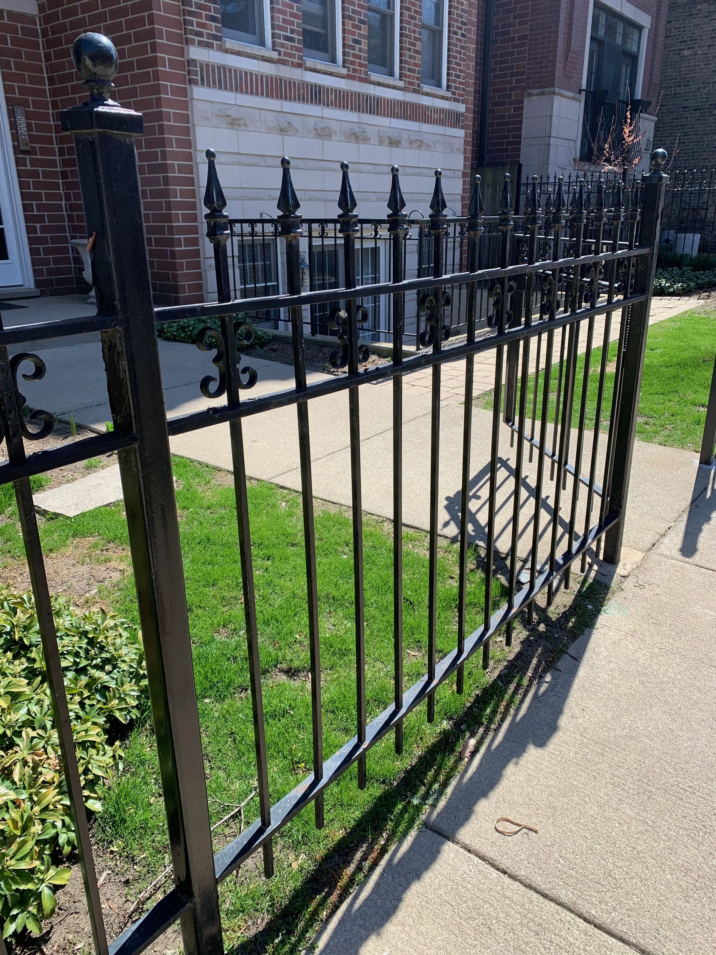Black iron fence with decorative finials surrounds a small patch of grass and sidewalk in front of a brown brick building.