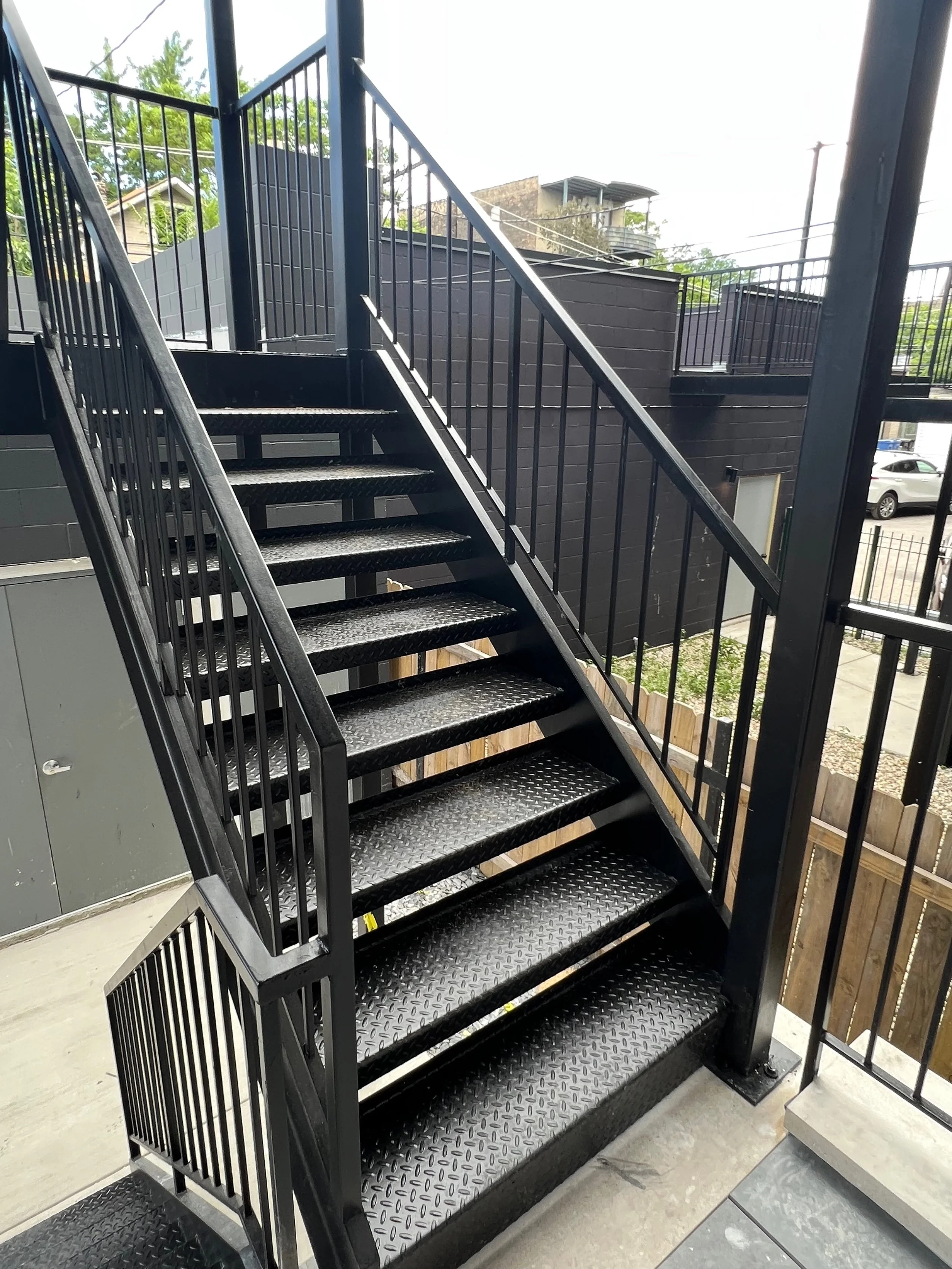 Black metal staircase with diamond plate steps leading to an outdoor balcony with a metal railing, surrounded by a brick wall and neighboring buildings.