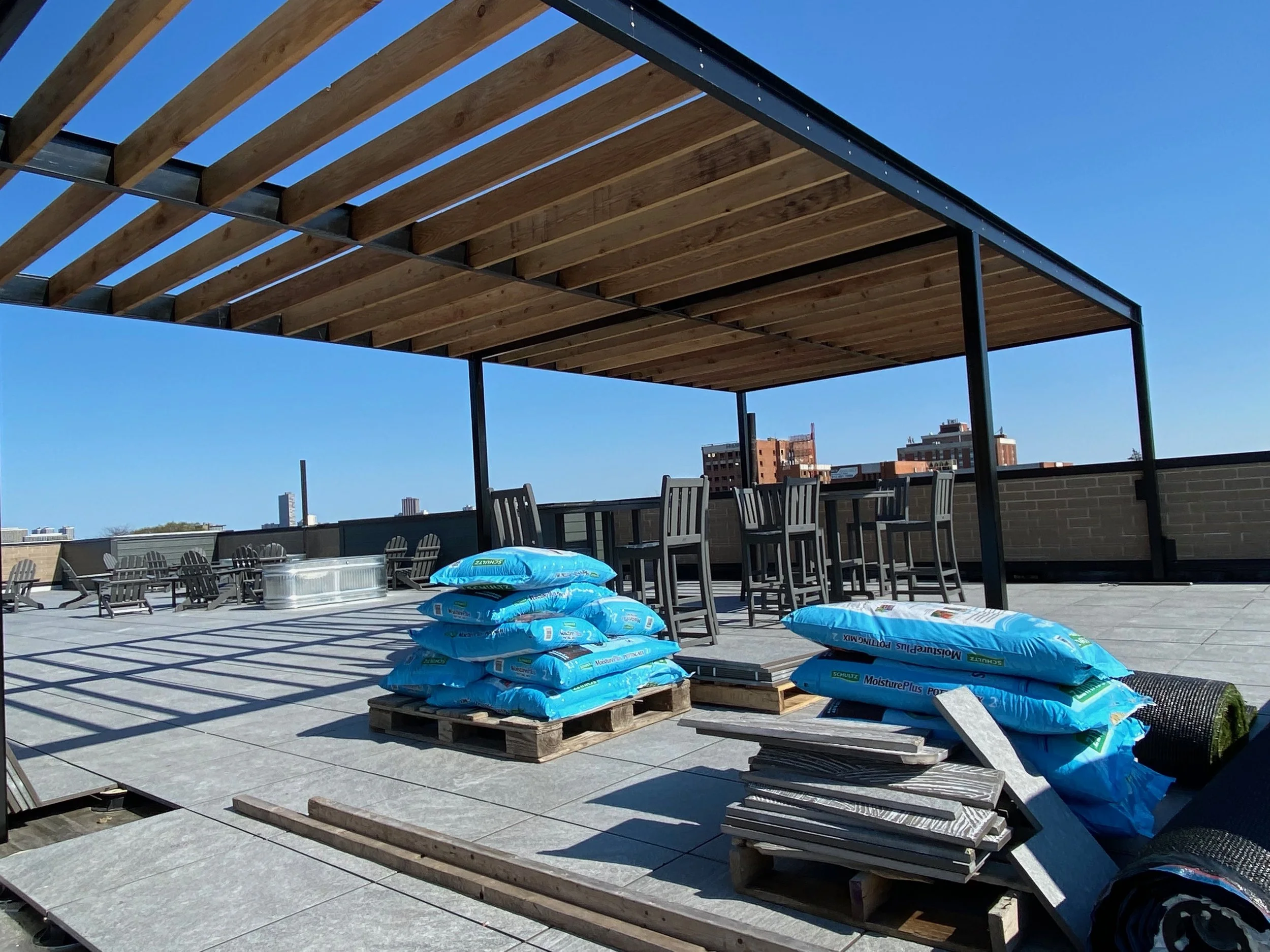 Construction site on a rooftop with stacks of bricks, bags of moisture protection materials, and outdoor furniture under a partially built wooden roof structure against a clear blue sky.