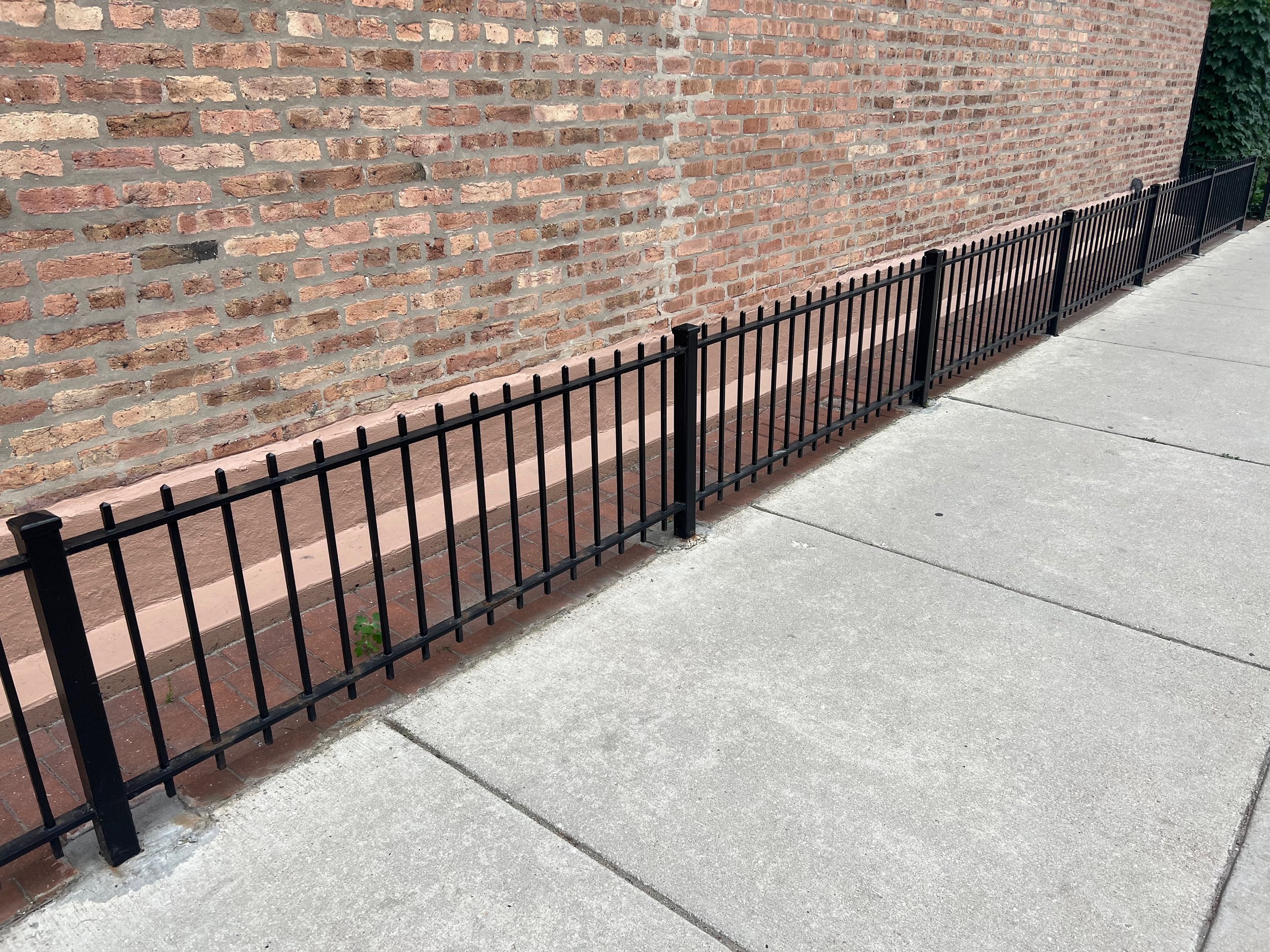 A red brick wall with a black metal fence running along the concrete sidewalk in front of it.