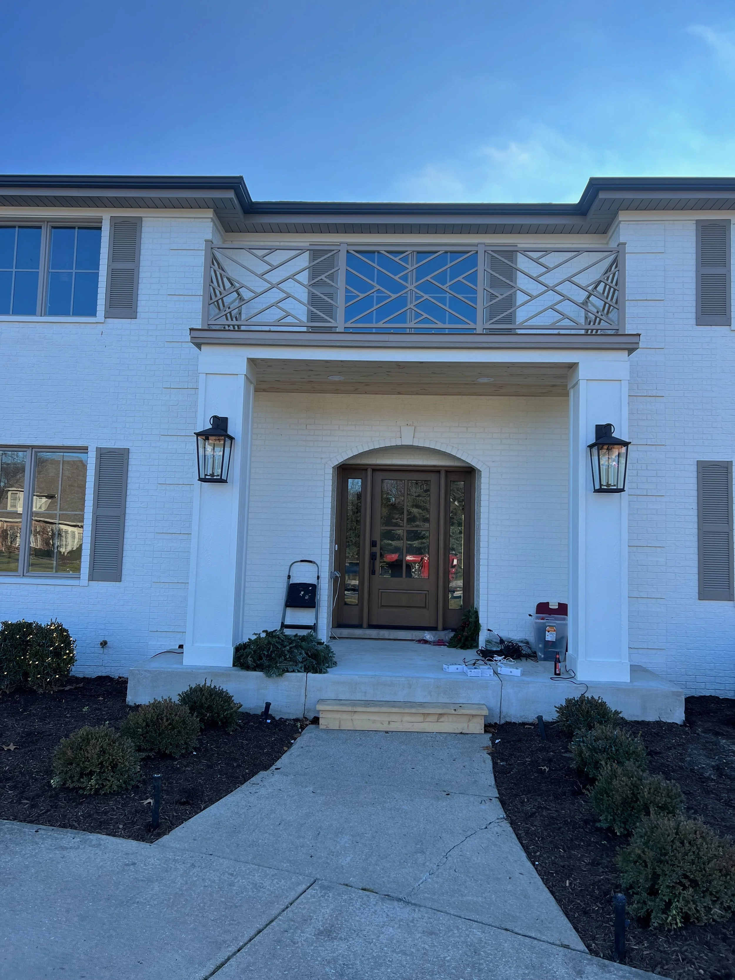 Front view of a white brick house with a covered porch, two black wall-mounted lanterns, a small set of steps, and some landscaping with small bushes. The house has a balcony with a geometric metal railing and large windows, with a partly cloudy sky 