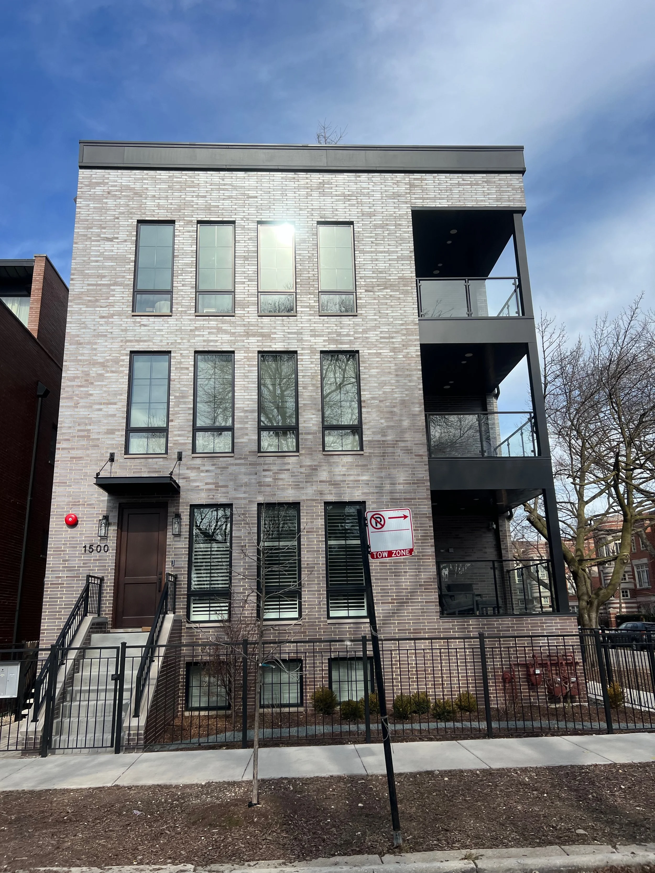 A modern multi-story brick residential building with black-framed windows and balconies, an entrance with stairs, a 'No Parking' sign, a red fire alarm bell, a small tree, and a sidewalk with a fence and some bushes outside. The sky is partly cloudy.
