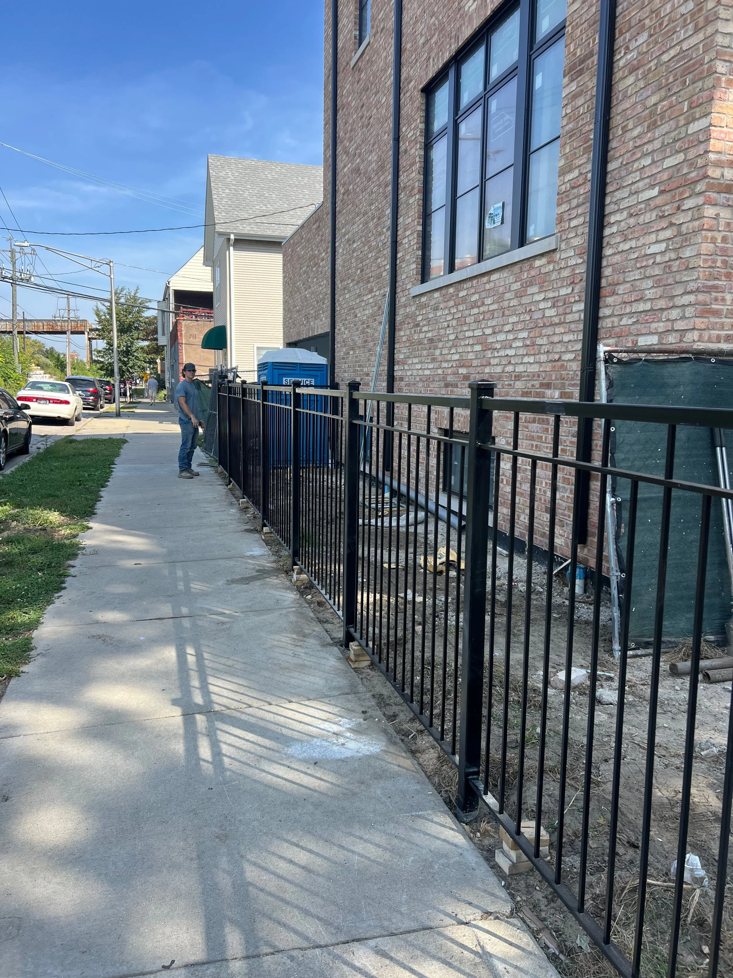 Sidewalk and residential building under construction with a black metal fence, a man standing on the sidewalk, power lines, parked cars, and a partly cloudy sky.