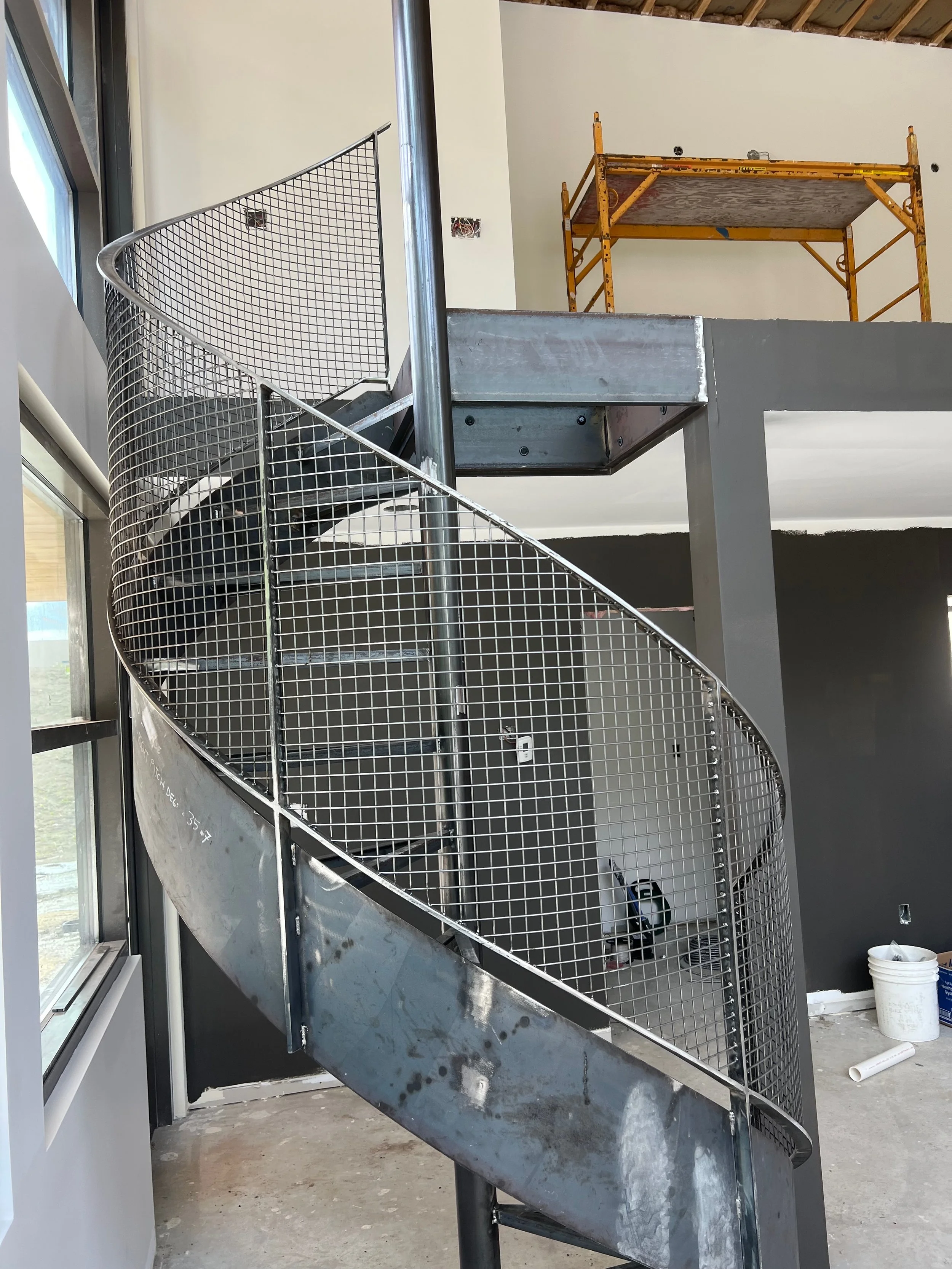 Stainless steel spiral staircase under construction inside a building, with a metal safety mesh on the outer side and construction scaffolding visible in the background.