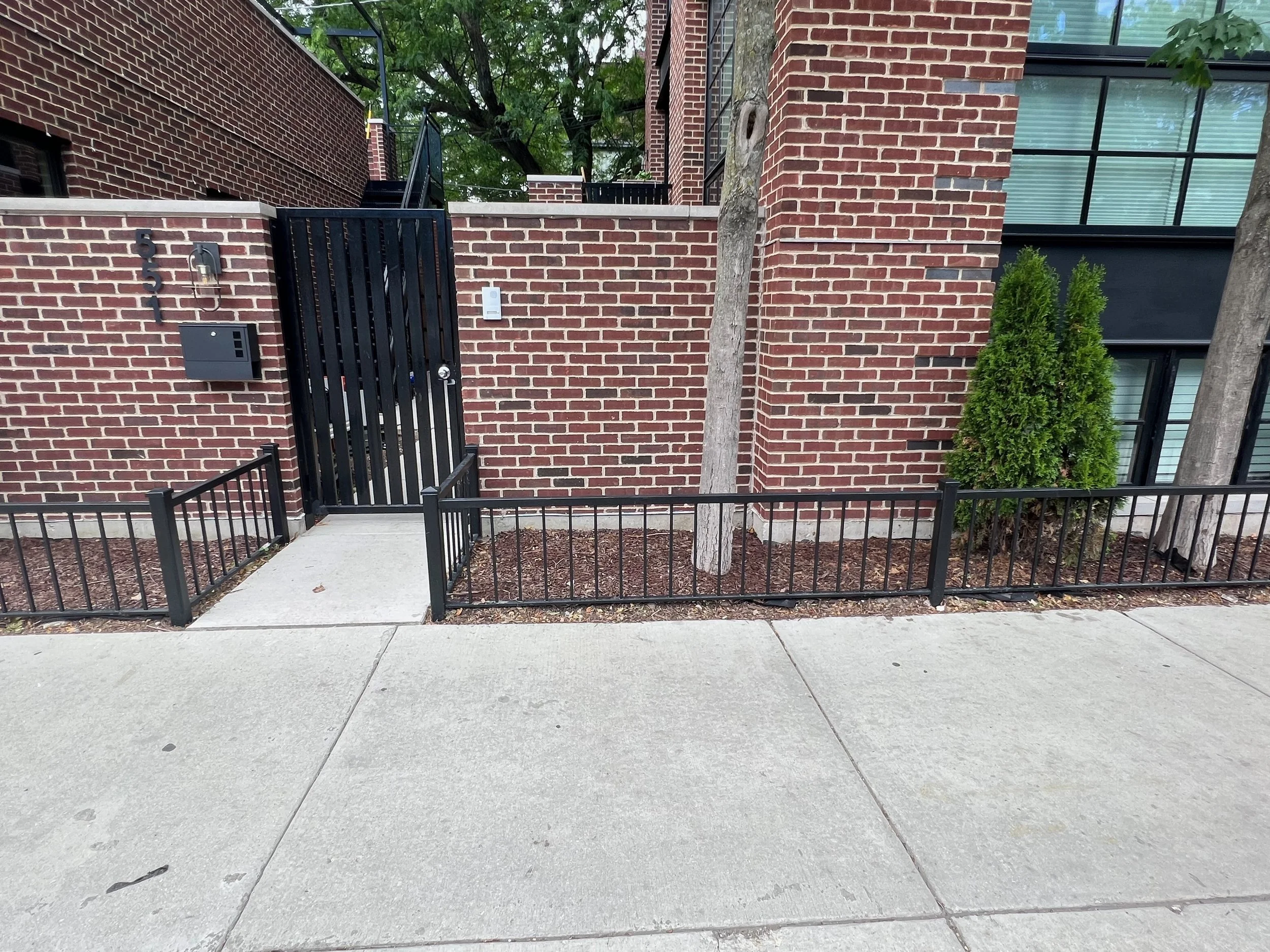 A residential building with a brick wall, a black metal gate, and stairs leading to upper floors. There are two trees in front, a small shrub, and a concrete sidewalk.
