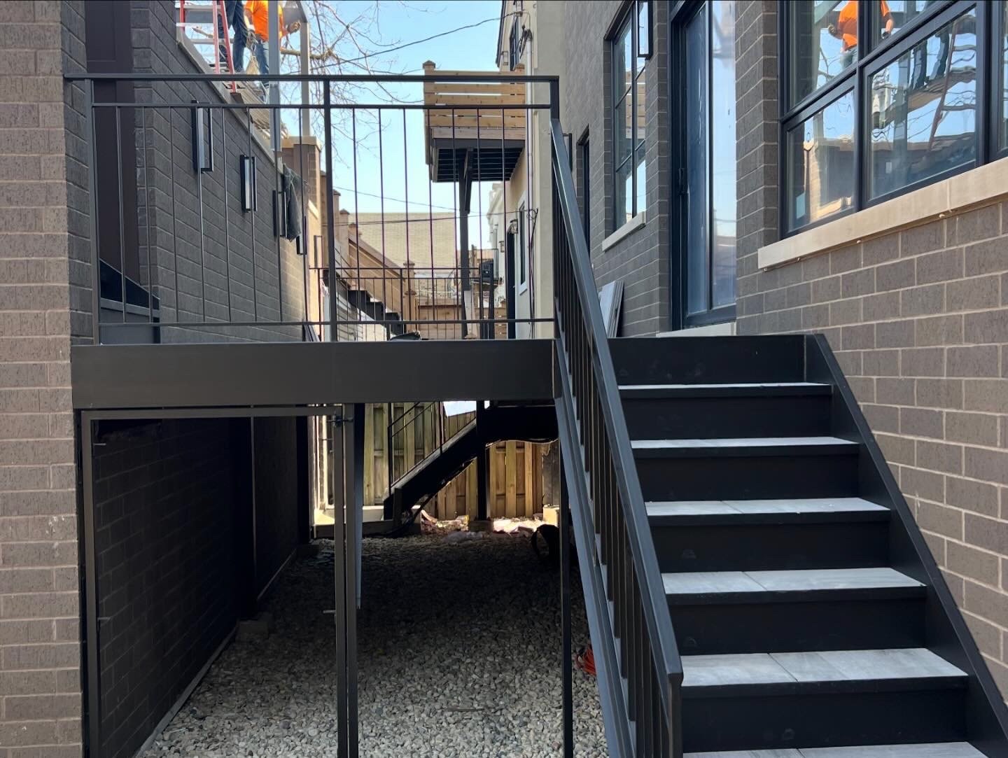 Backyard outdoor stairs and deck of a modern house with gray brick walls, black railings, and a gravel ground.
