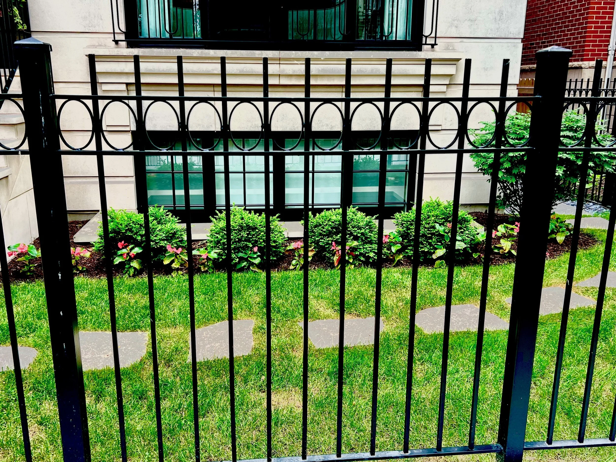 View of a small garden with green bushes and pink flowers behind a black iron fence in front of a building with beige and red brick walls and a glass window.