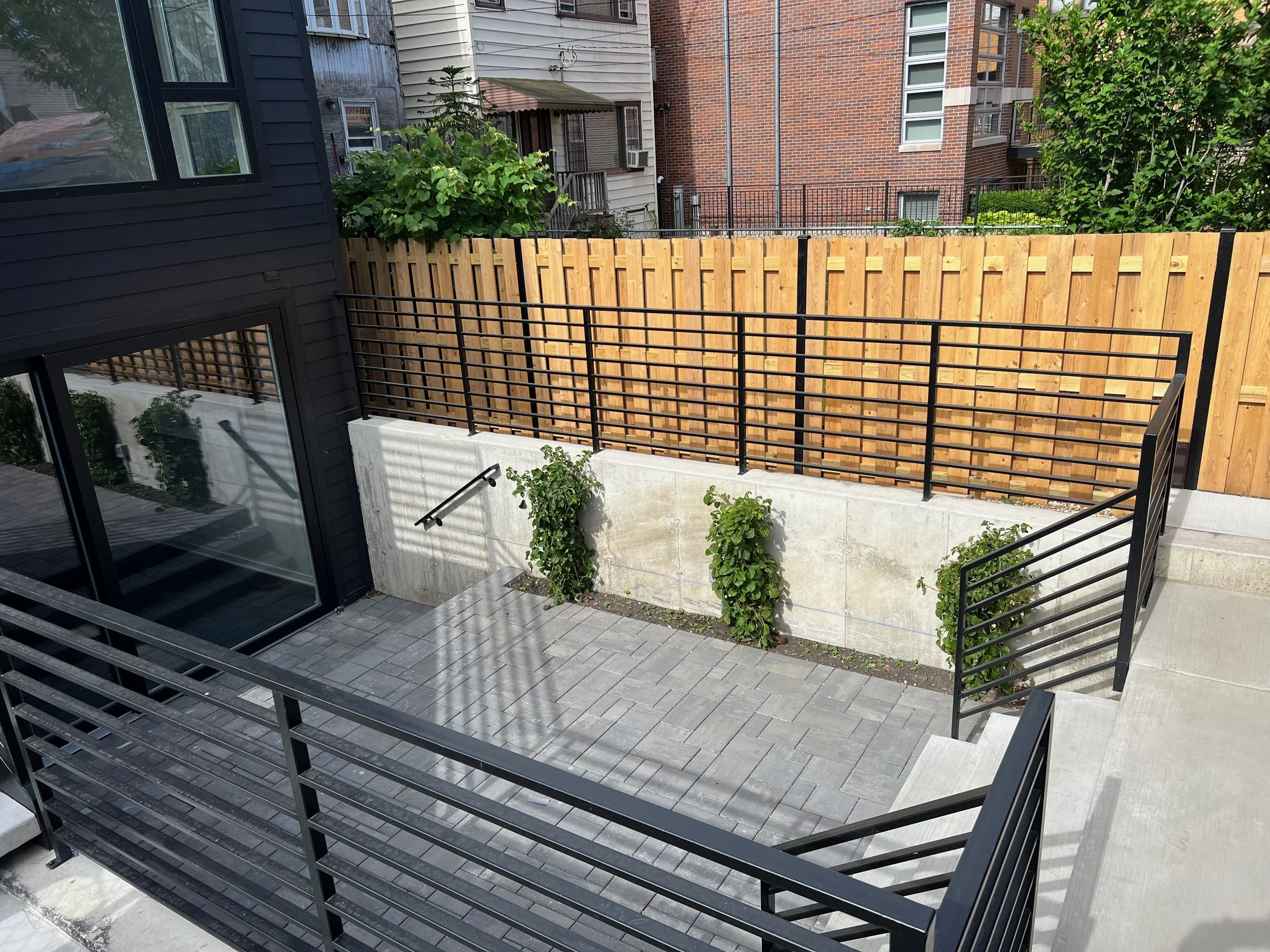 A residential outdoor patio area with a black metal staircase and railing, a wooden privacy fence, and neighboring brick and siding apartment buildings in the background.