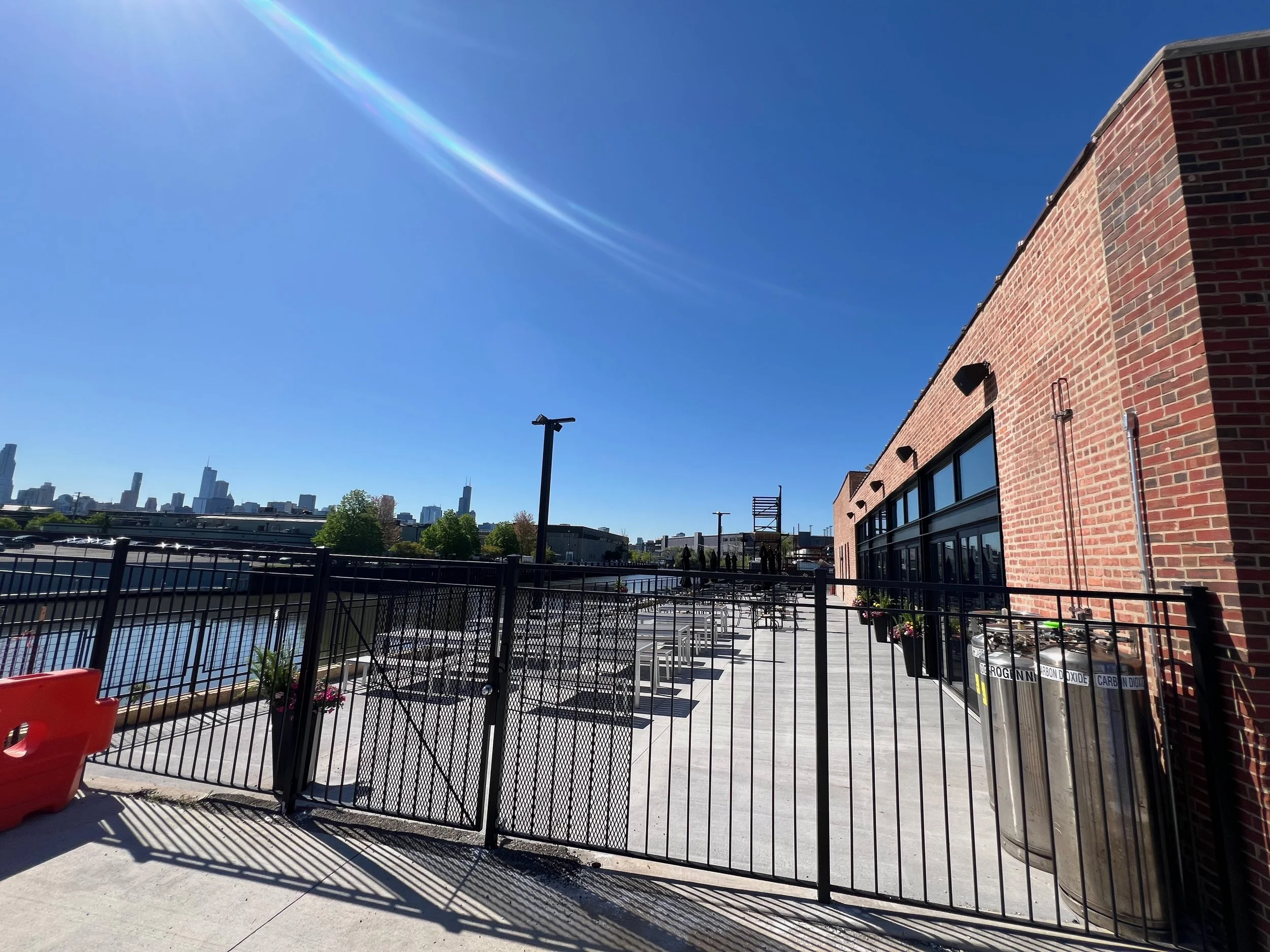 View of a rooftop patio area with black metal fence, brick building wall, city skyline in the background, and clear blue sky with sunlight.