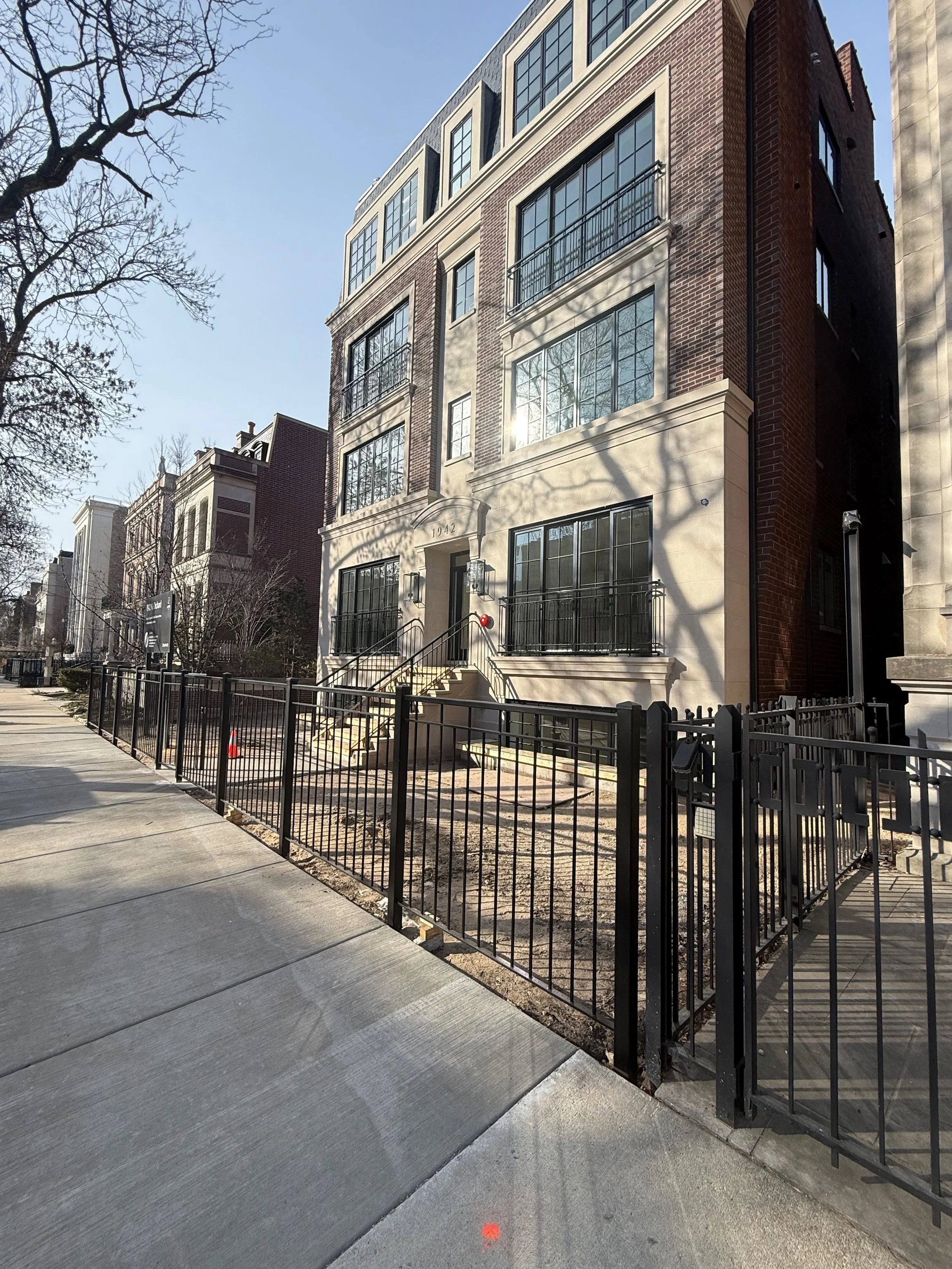 A multi-story residential building with large windows, brick and beige-colored exterior, and small balconies. The building is located on a city street with a sidewalk, a black metal fence, and some leafless trees.