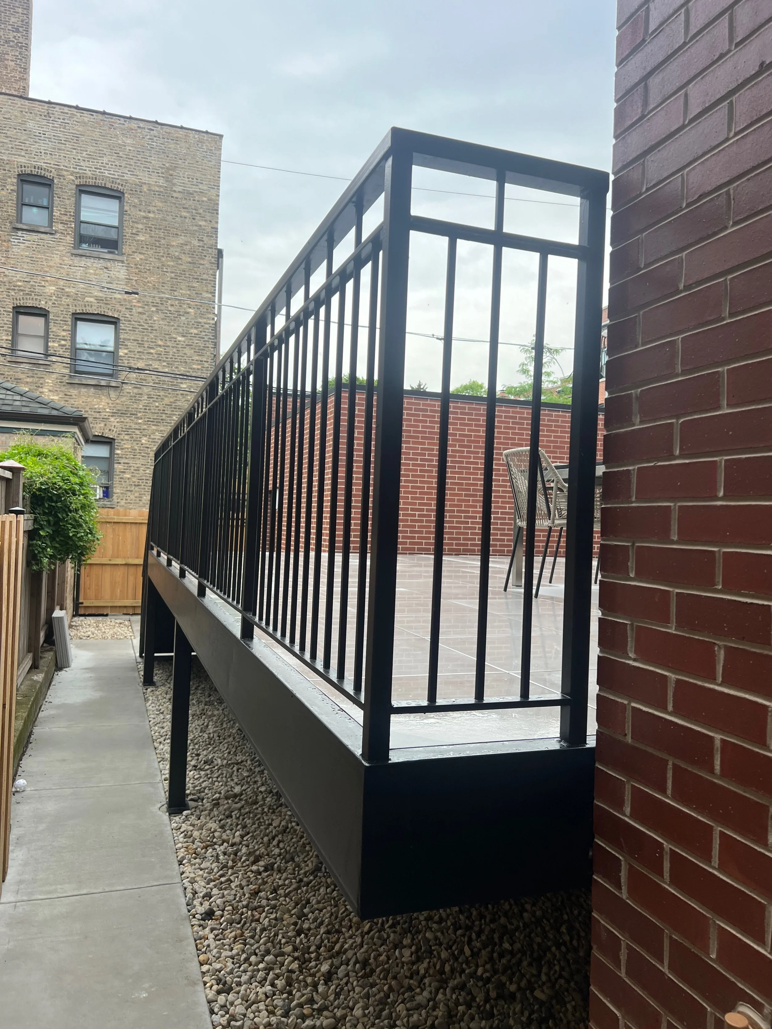 A small outdoor patio area with a black metal railing, red brick wall, and dark bricks flooring. The patio is adjacent to a building with a brick wall and a garden area with a wooden fence and some greenery. Overcast sky in the background.