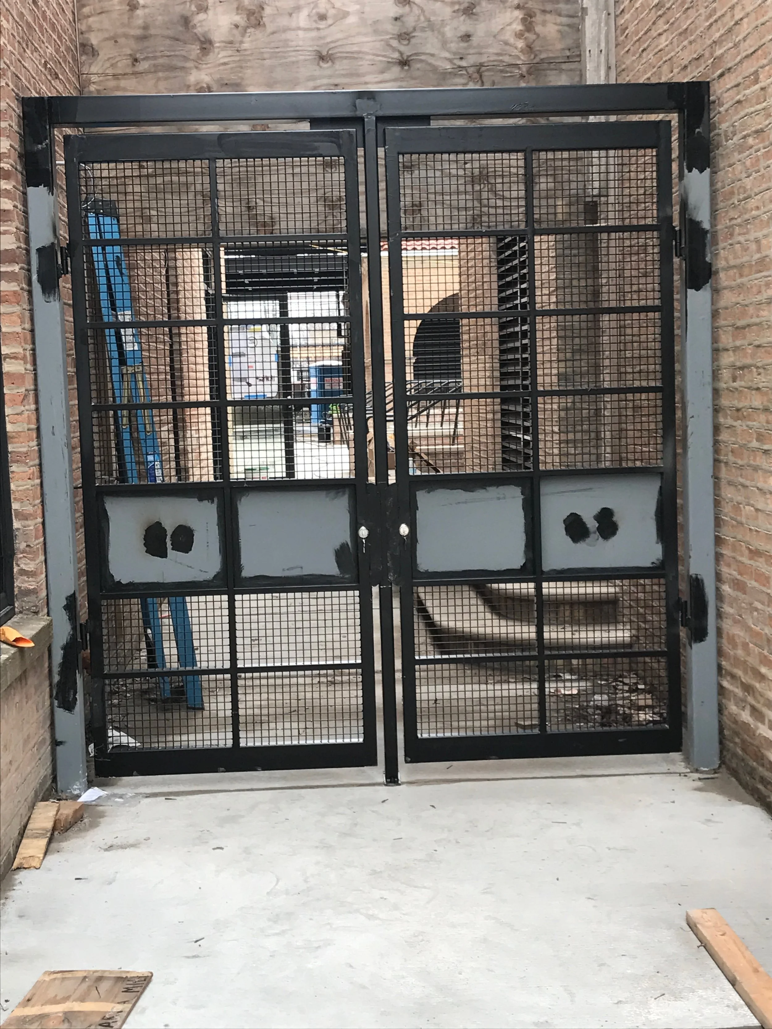 Black metal gate with mesh design, in an enclosed outdoor area with brick walls and concrete floor, construction materials around.