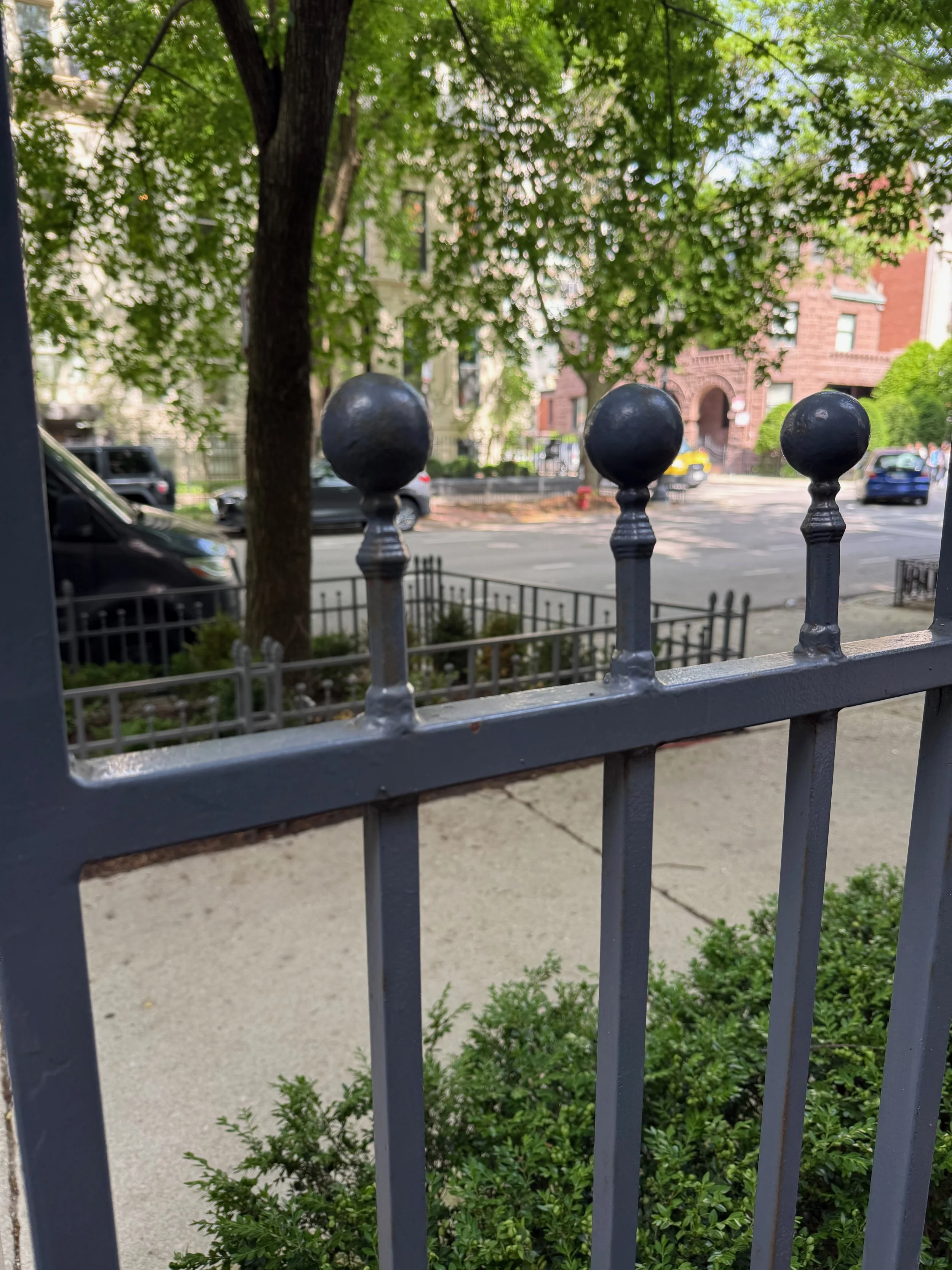 View of a city street taken from behind a black metal fence with decorative finials. In the background, there are parked cars, a sidewalk, trees with green leaves, and buildings made of brick and stone.