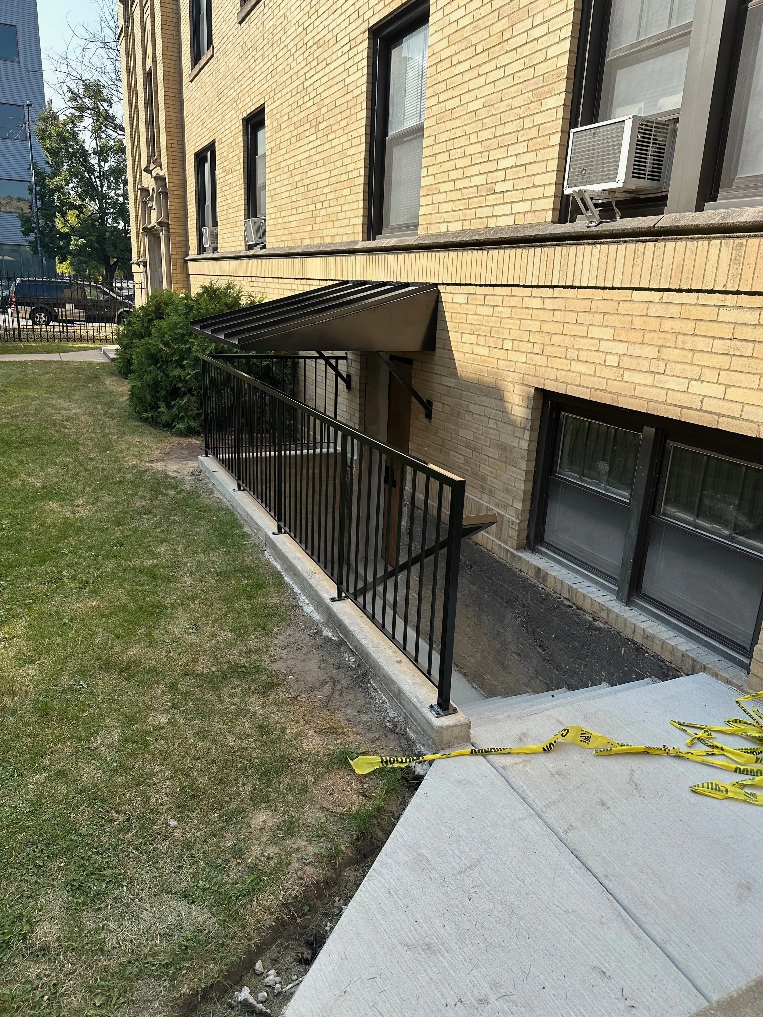 Construction site outside a brick apartment building with newly installed black metal railing along concrete stairs and partially replaced concrete sidewalk, caution tape, green bushes, and parked cars in the background.