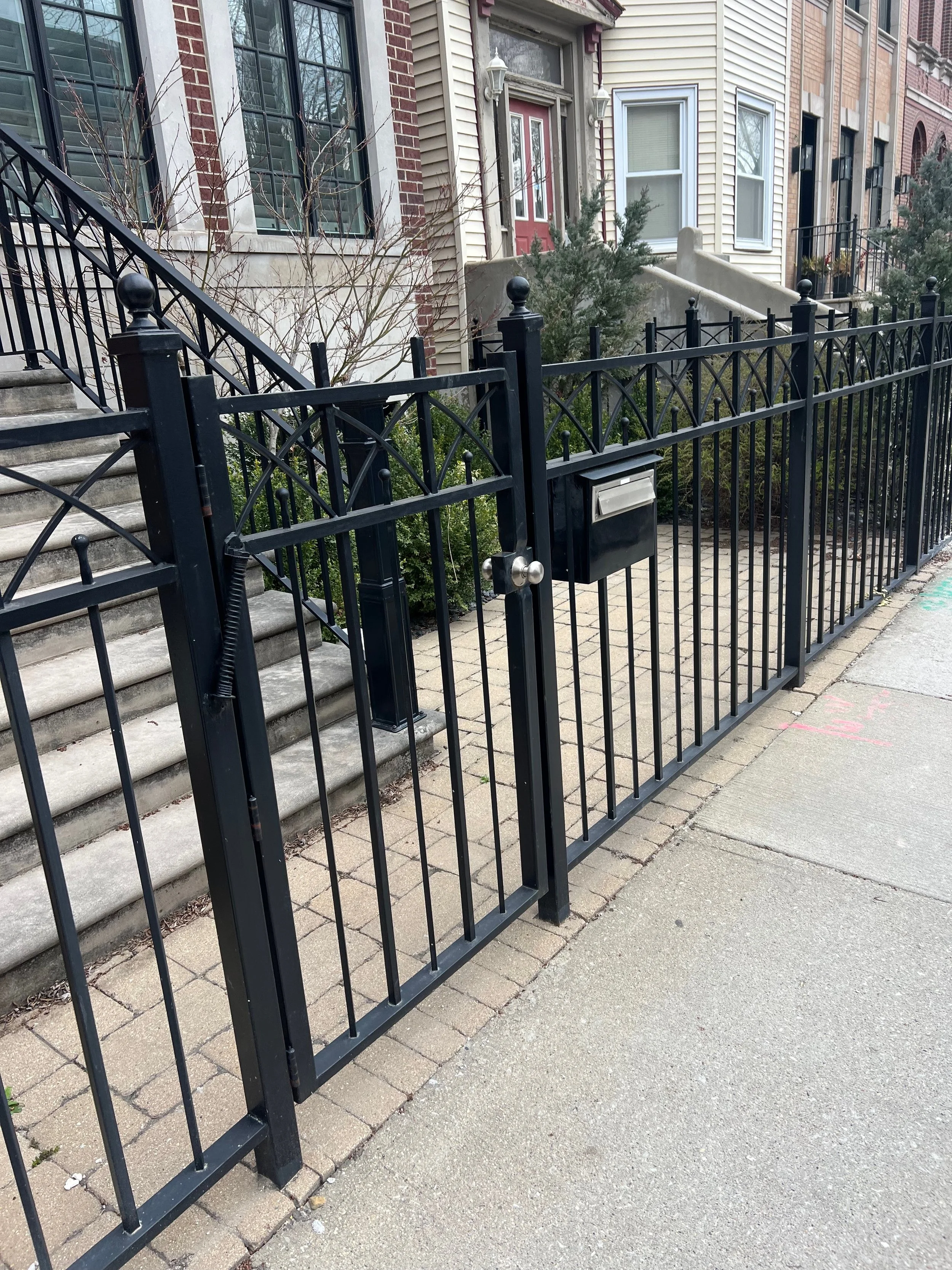 Front yard of row houses with black metal fence, concrete stairs, shrubs, and a mailbox.