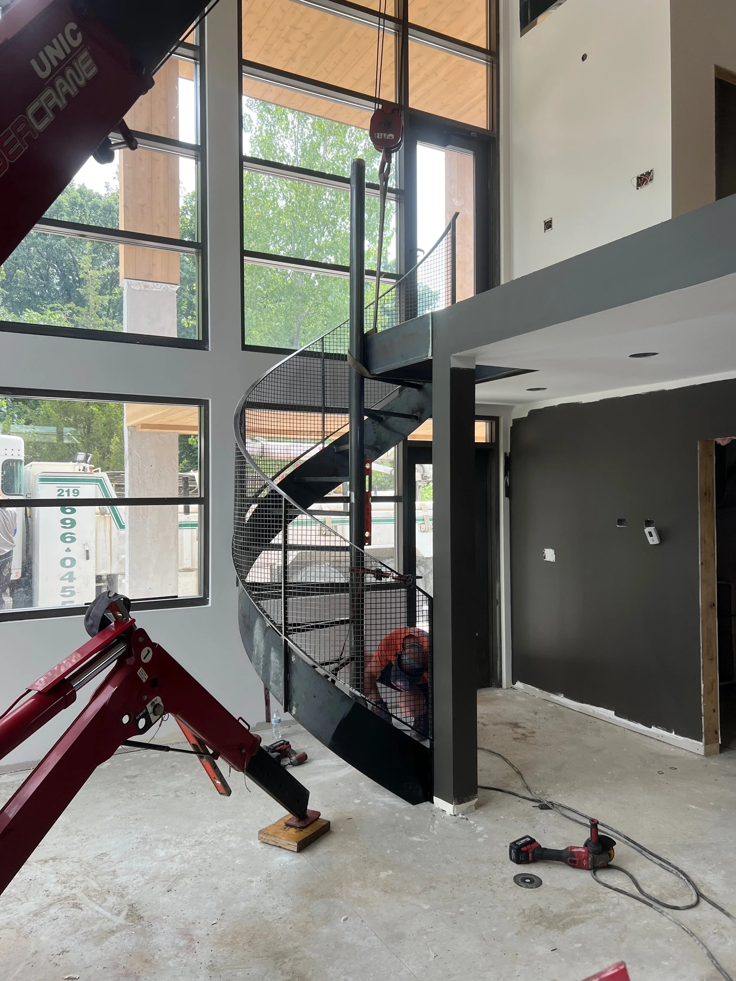 Interior of a building under construction with a spiral staircase, large windows, and construction tools and equipment on the floor.
