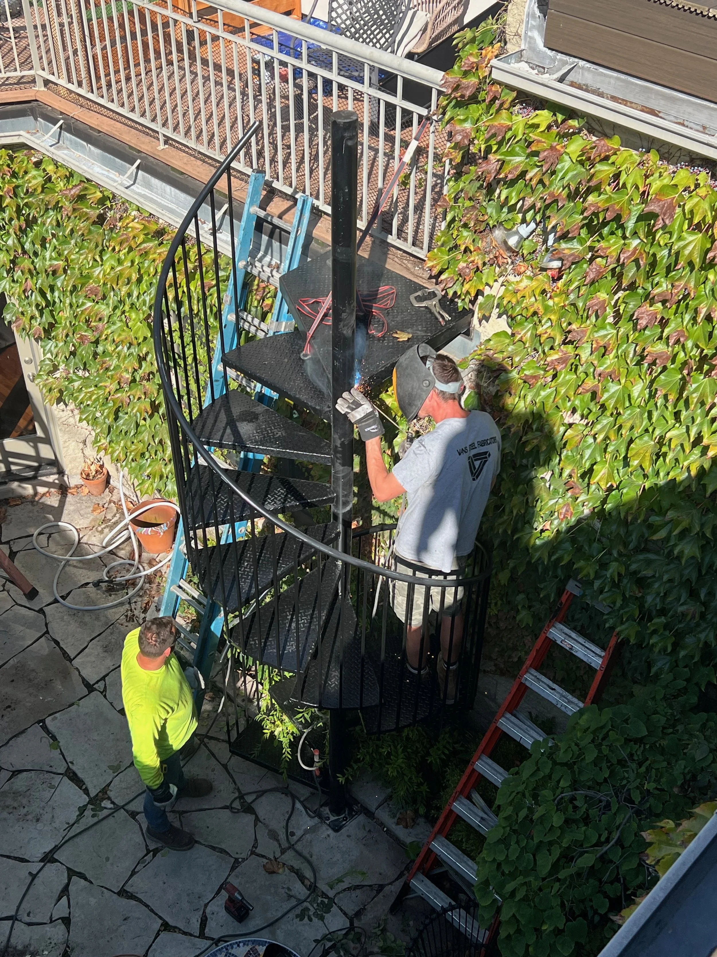 Construction workers assembling or installing a spiral staircase outdoors, with one person on the staircase, another standing below, and a third working on the scaffolding nearby.