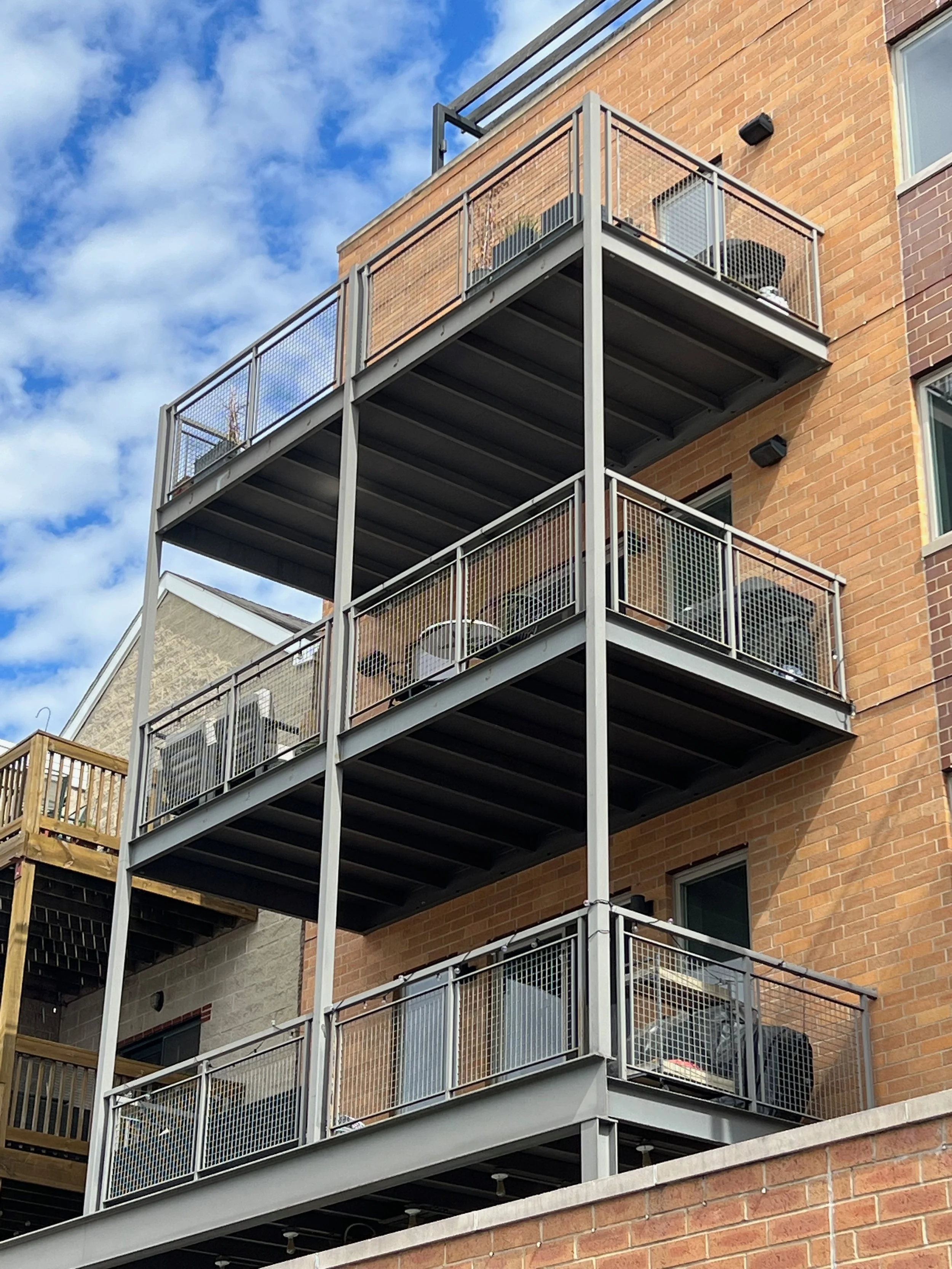 Multiple residential apartment balconies with metal railings on a brick building against a background of blue sky and clouds.