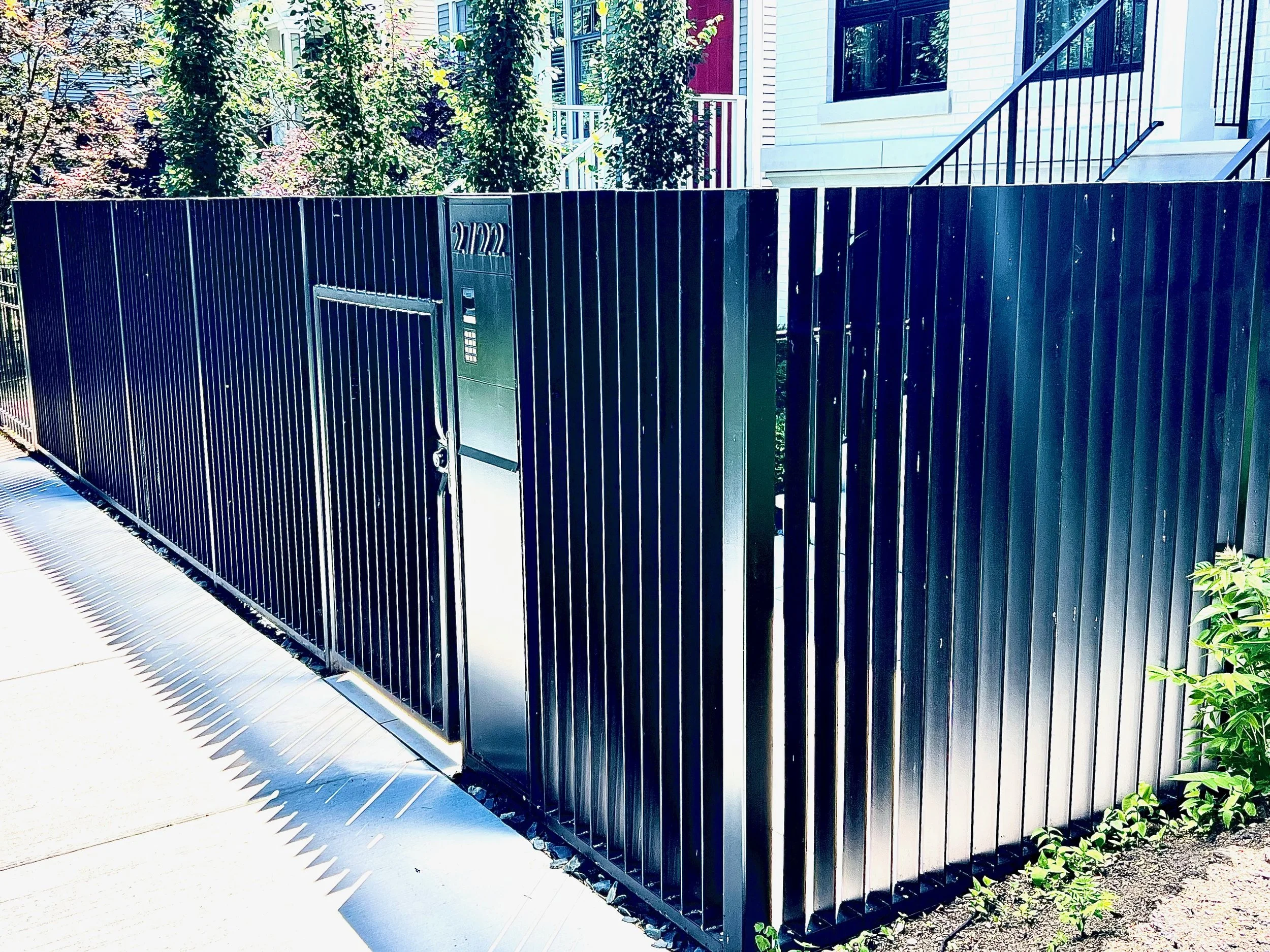 A modern black metal fence with a built-in mailbox and keypad, enclosing a residential area with townhouse-style homes and green trees in the background.