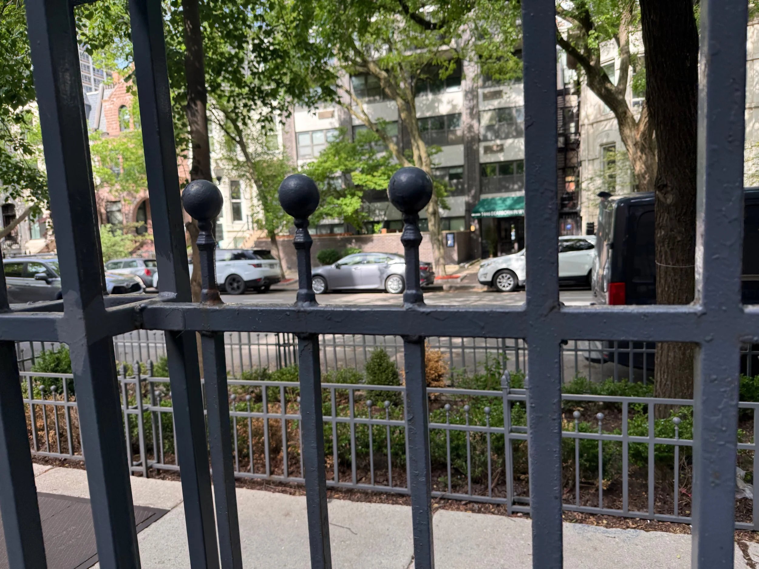 View of a city street seen through black metal fence with decorative round finials. Behind the fence, there are parked cars, trees with green leaves, and apartment buildings in the background.