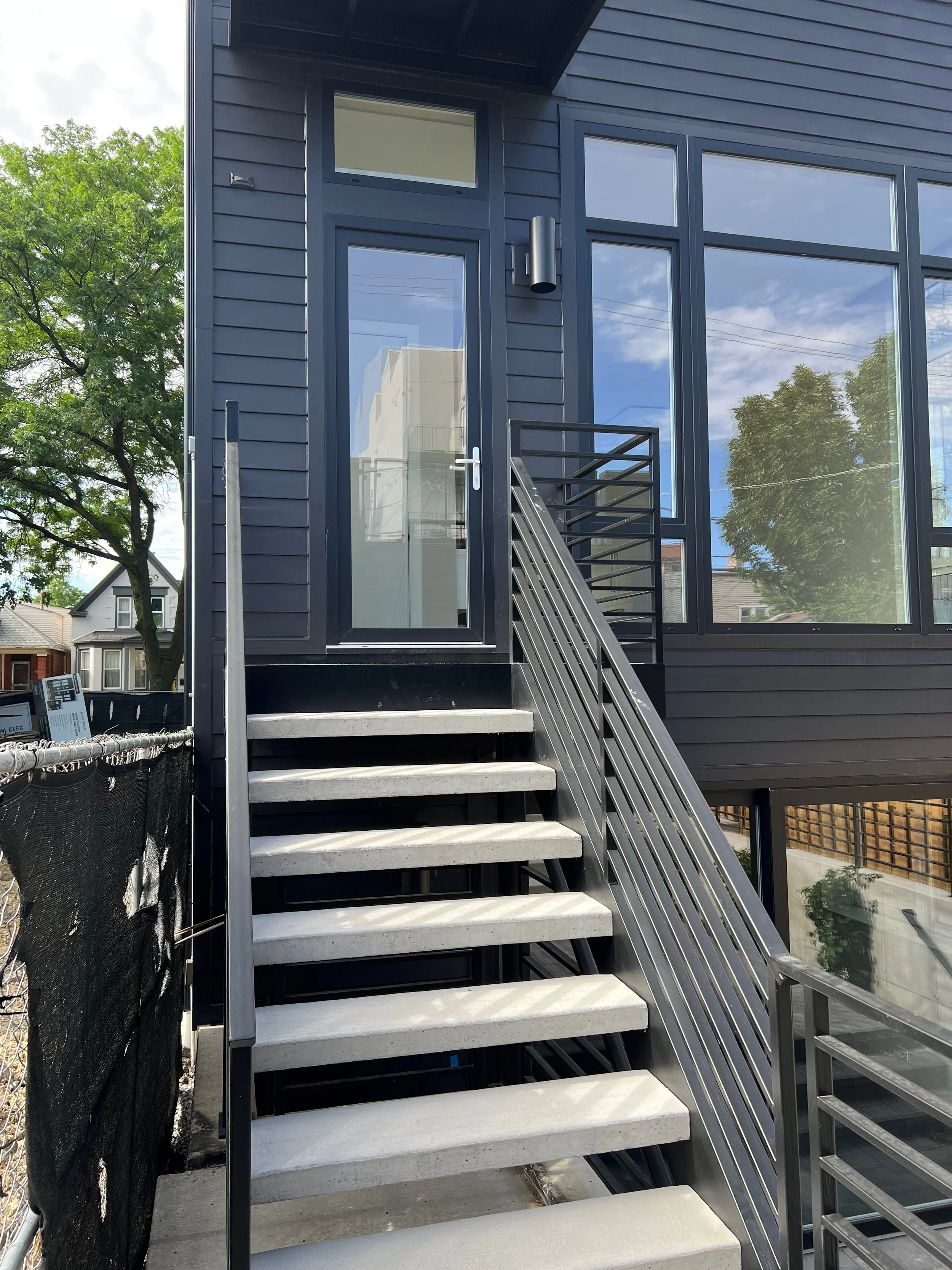 Exterior staircase with concrete steps, metal railings, outside modern black building with large glass windows, trees, and neighboring houses