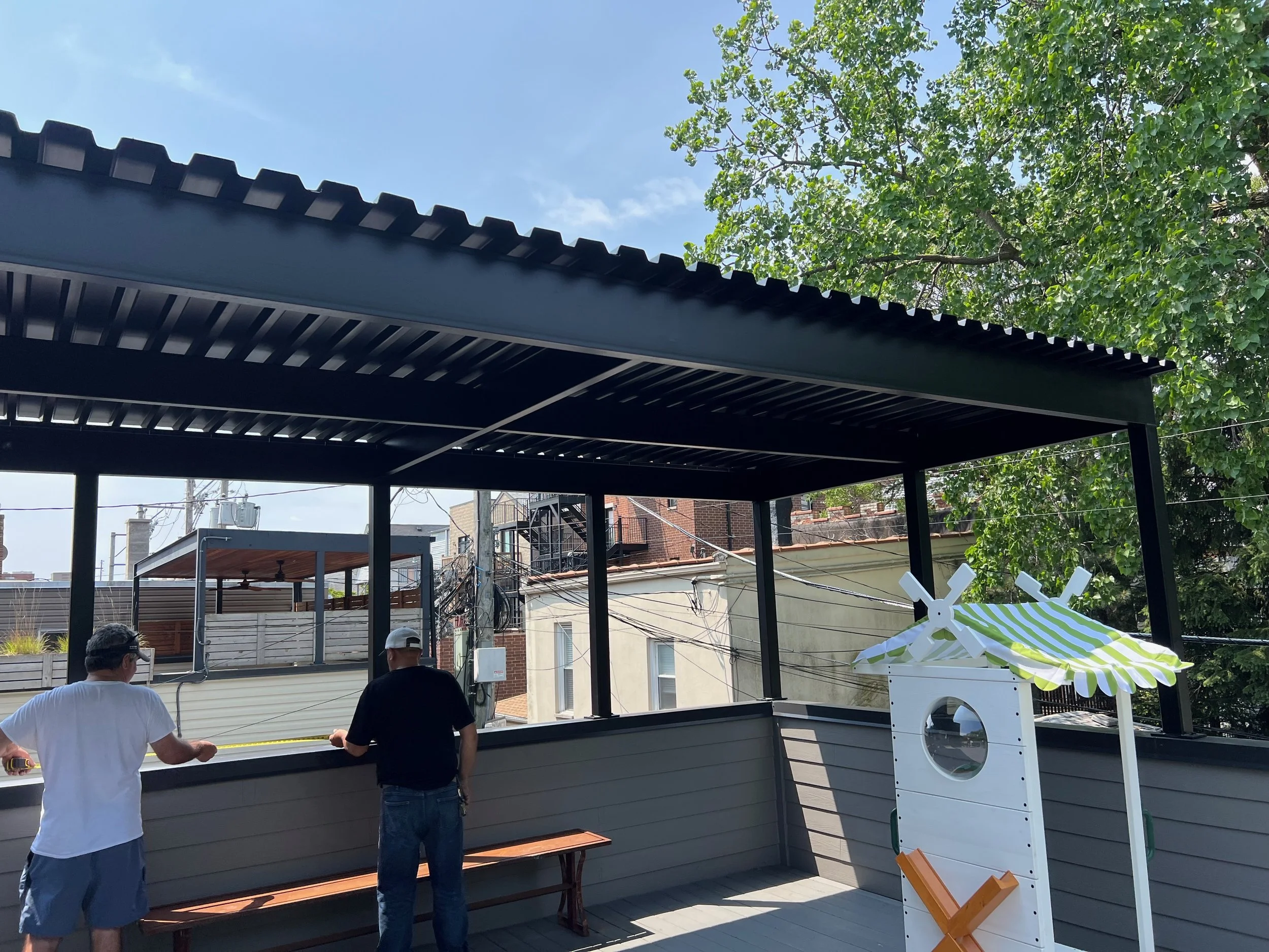 Two men standing in an outdoor rooftop area with a black bar counter, some wood benches, and a white washing machine with a green and white striped umbrella.