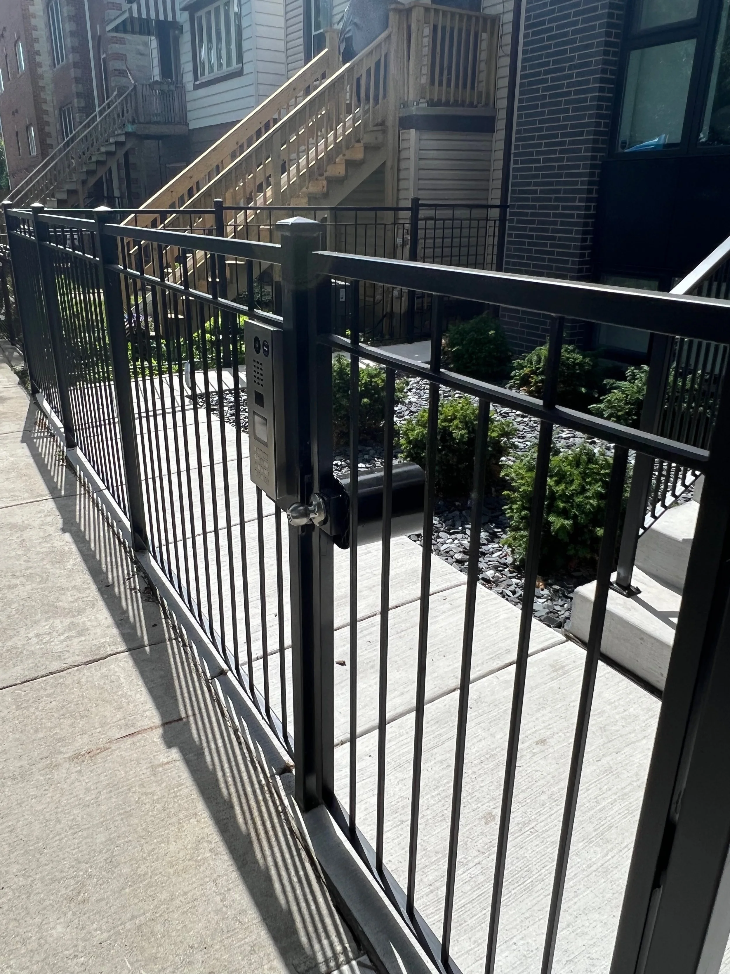 A fenced patio area with stairs leading up to a building entrance, with a keypad lock on the gate and some plants inside the fenced area.