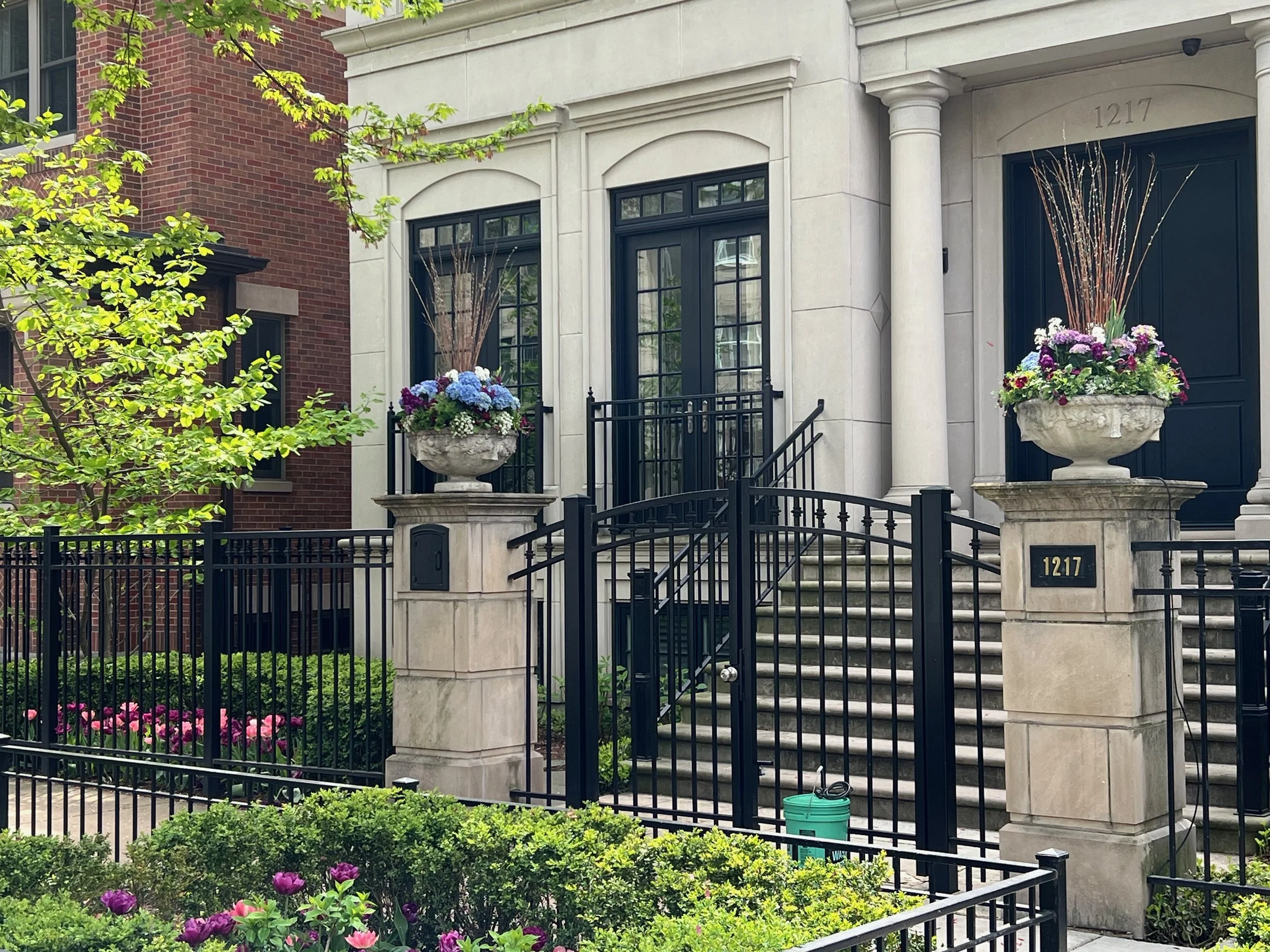 View of the front entrance of a residential building with stairs, black iron gate, large planters with purple and white flowers, and green foliage in the foreground. The building has a beige stone facade with black framed windows and brick on the sid