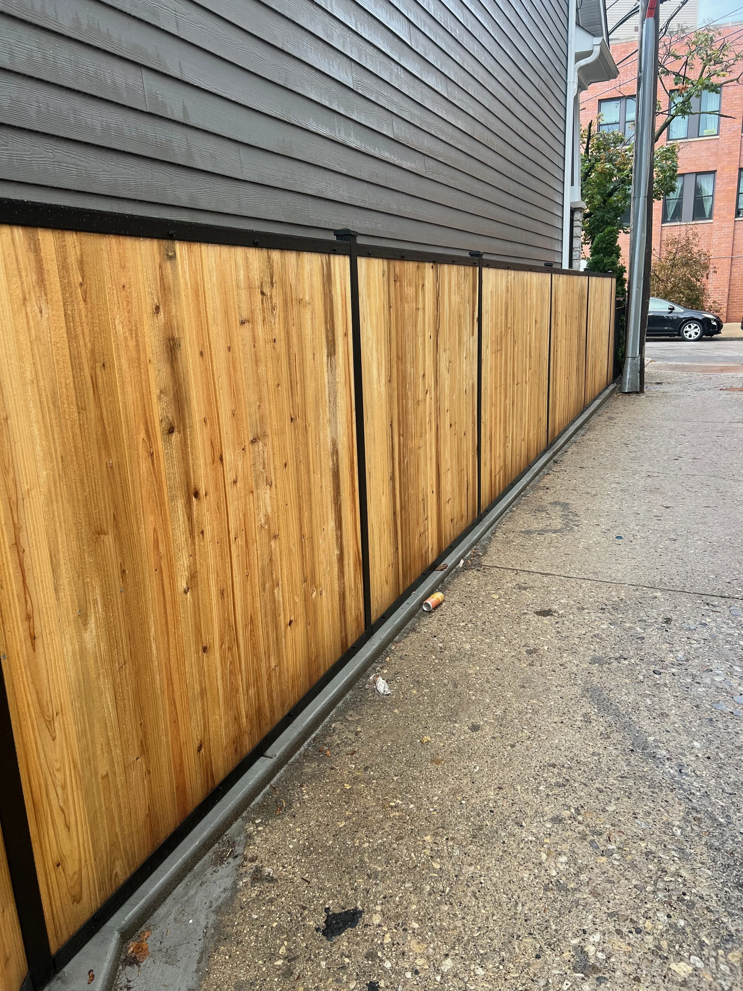 Wooden fence with black metal framing alongside a building with gray siding, on a concrete and dirt sidewalk in an urban area with a parked car and some trees in the background.