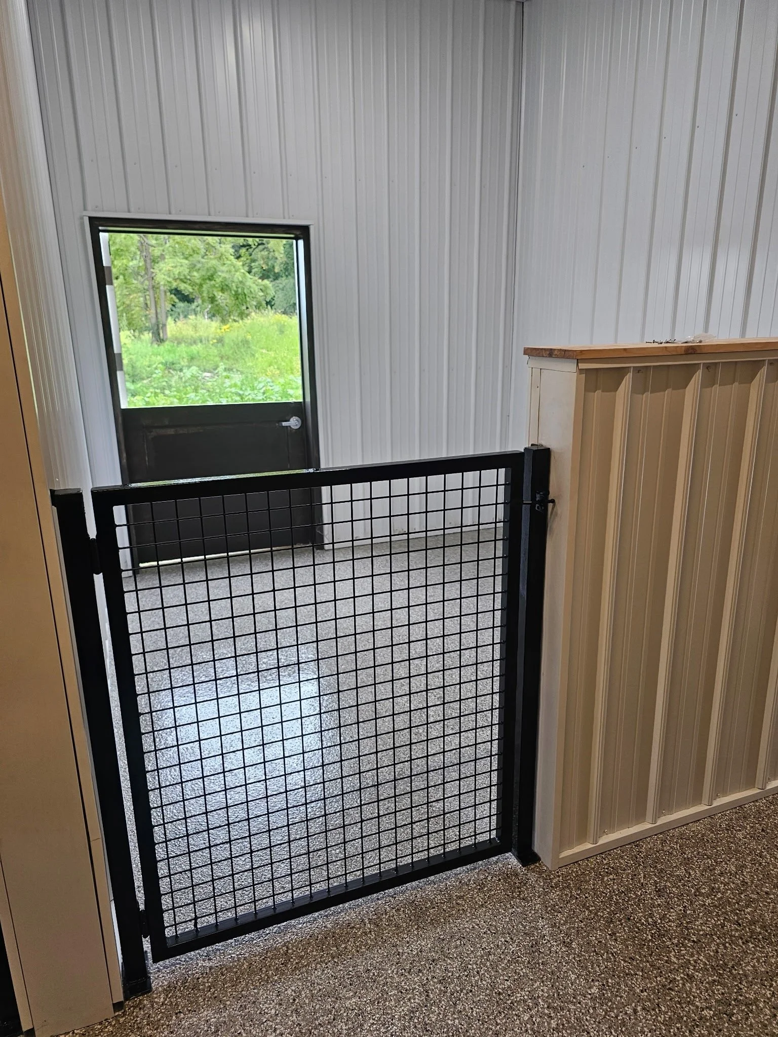 Interior view of a small enclosed porch or mudroom with a black metal gate, a window showing greenery outside, a partial view of a wooden panel wall, and a speckled carpeted floor.