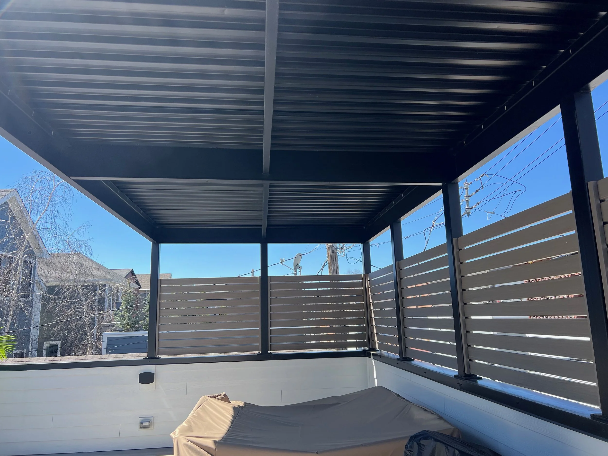 A view of a balcony or patio area with a metal roof, side walls with horizontal slats, and trees and houses visible in the background on a sunny day.