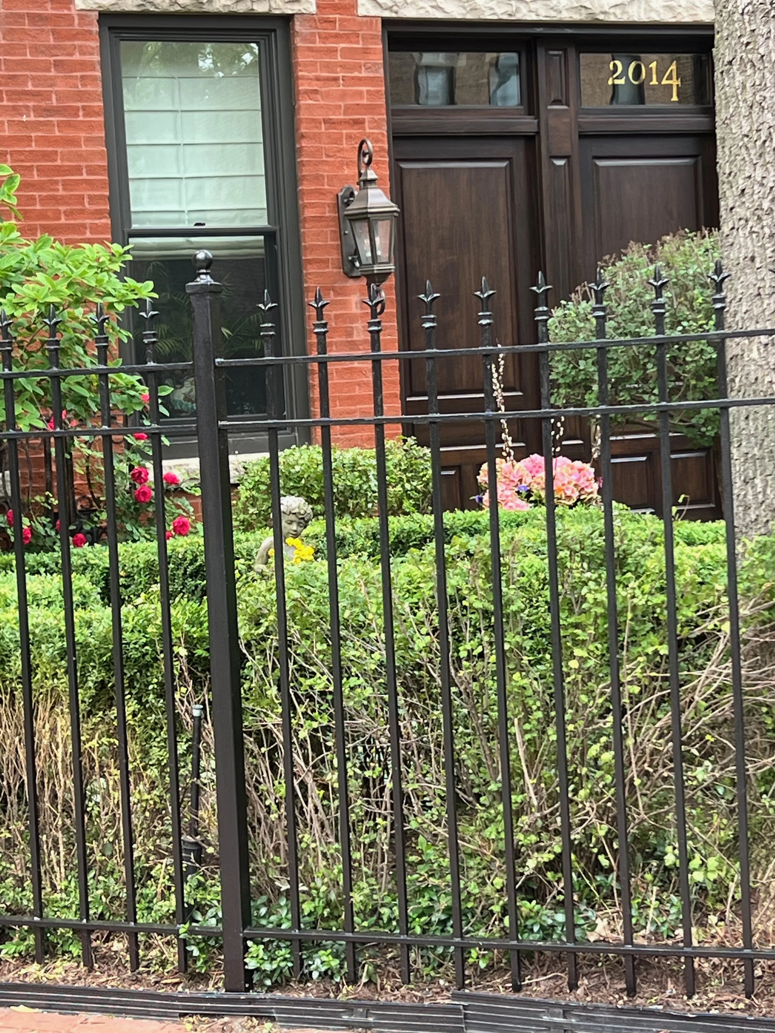 A house with a brick exterior and a black front gate. The house has a large window with the number 2014 above a dark wooden door. There is a porch light next to the window and many colorful flowers and shrubs in a garden in front of the house.