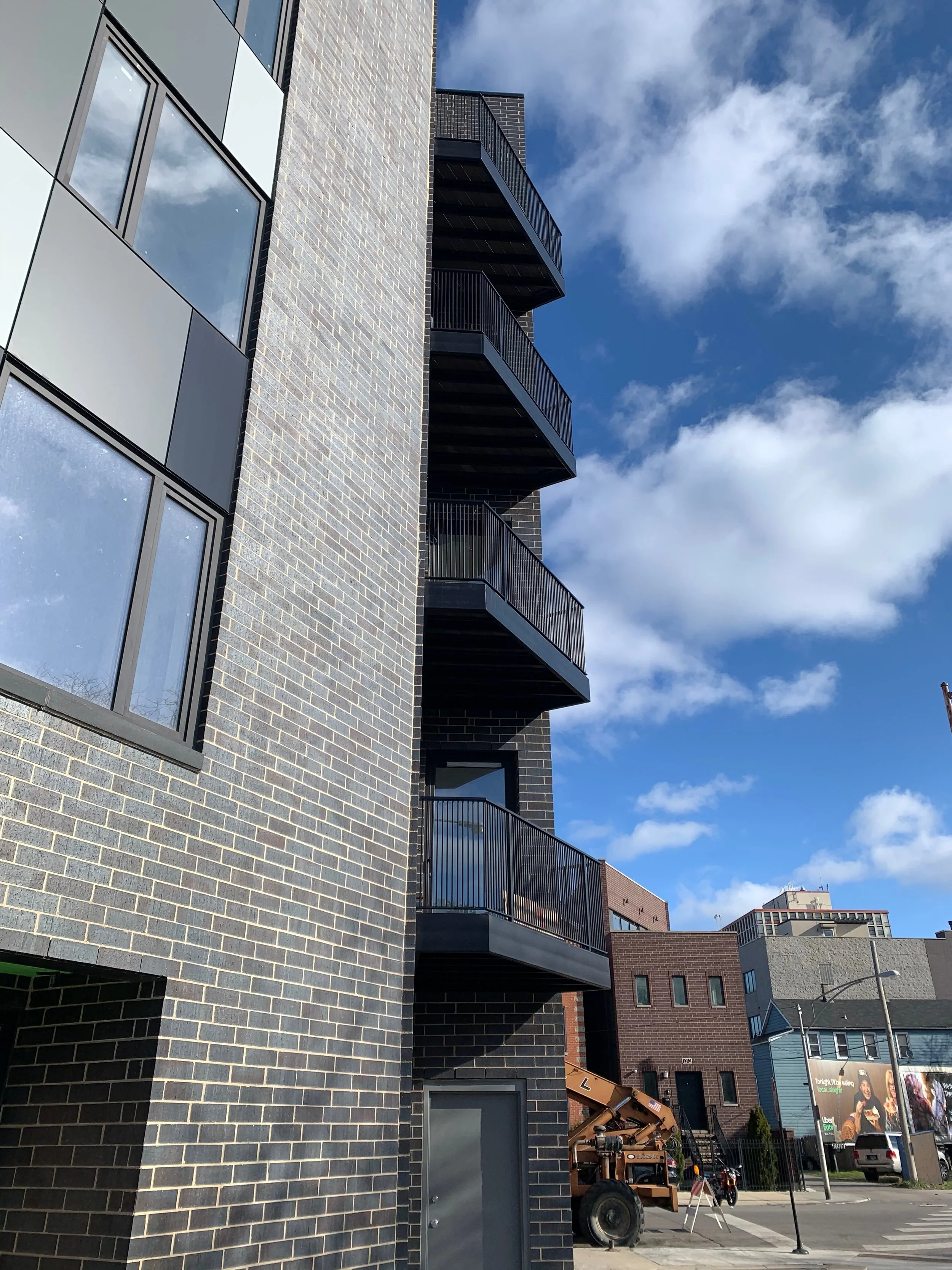 Modern multi-story apartment building with black brick exterior, balconies, large windows, and a blue sky with scattered clouds.