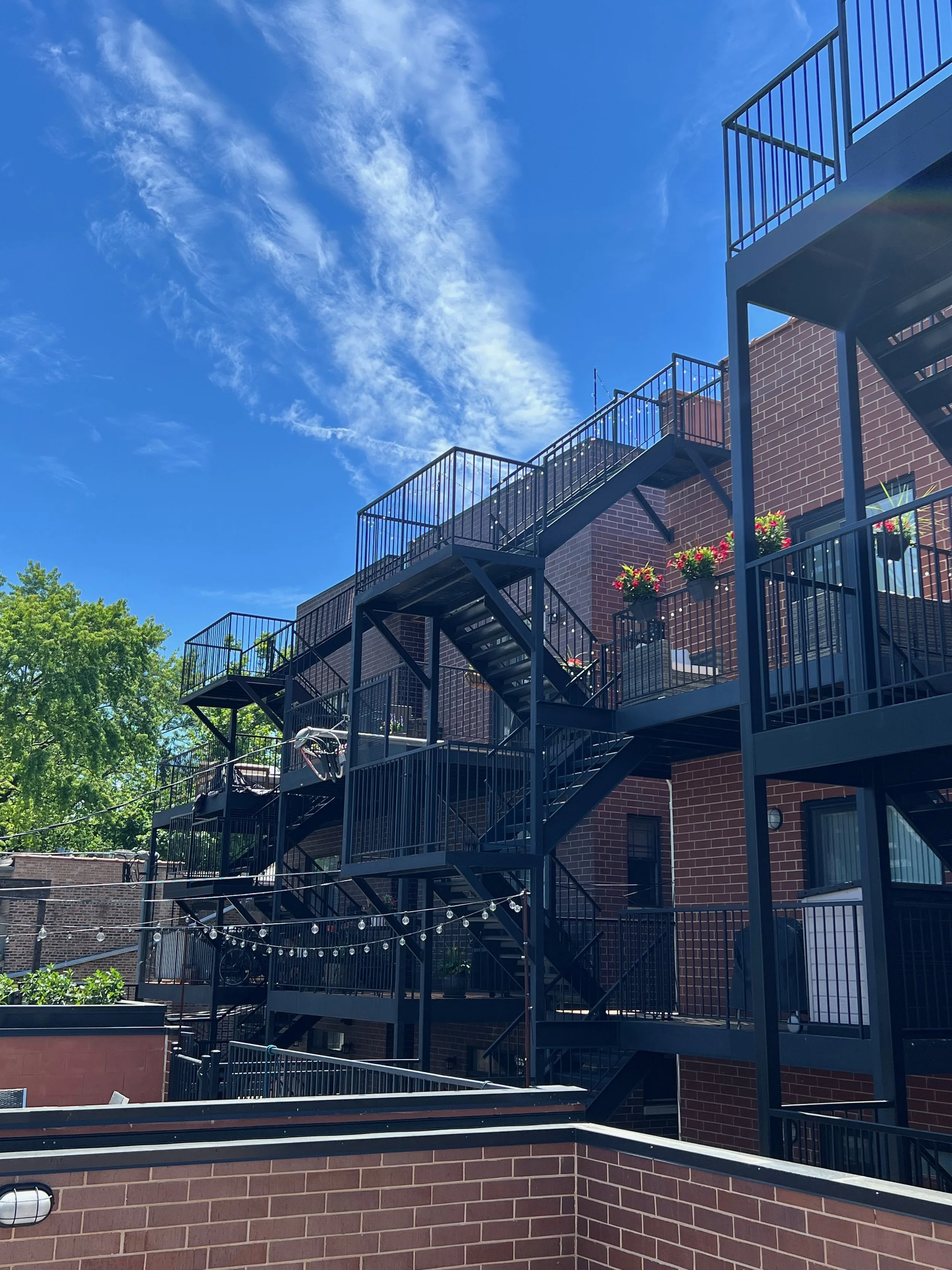 Multiple black fire escape stairs attached to the brick building with potted flowers on the balcony, against a clear blue sky with wispy clouds and green trees.